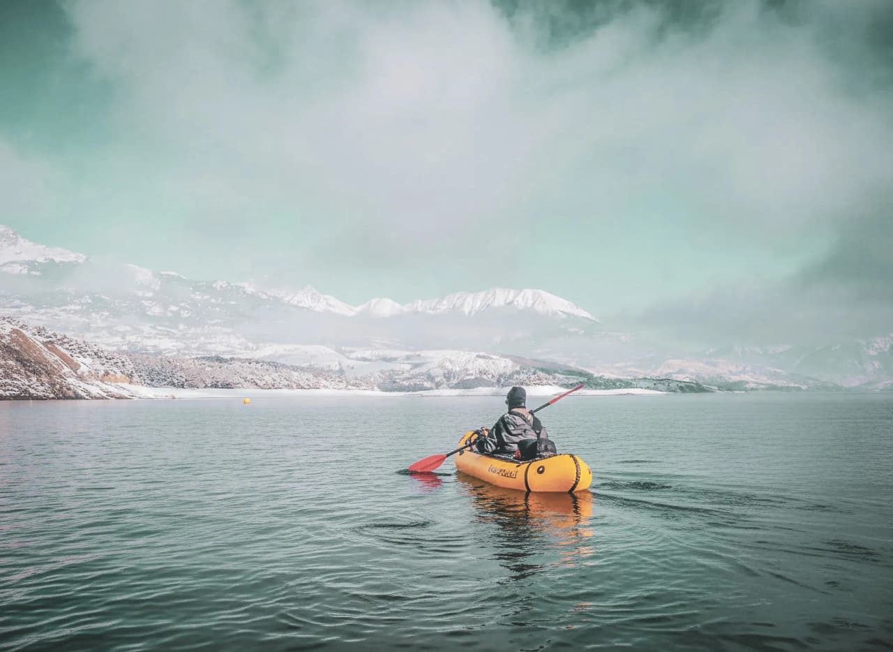 Un aventurier pagayant en packraft sur le lac de Serre-Ponçon, entouré de montagnes enneigées.