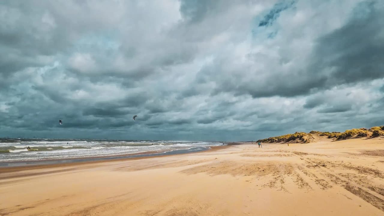 A golden beach fringed by dunes, under cloudy skies, with kitesurfers in the distance. Adventure in Zeeland!