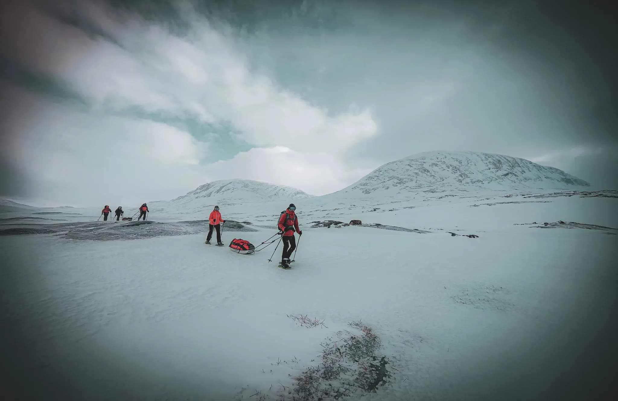 A group of people in ski gear, dragging pulkas across snow-covered terrain. They move together through a landscape of white mountains under a cloudy sky.