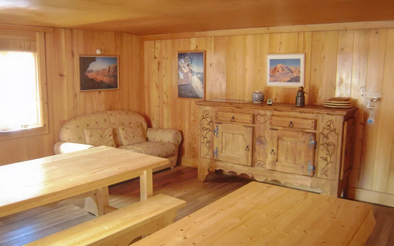 A warm wooden room, with light wood walls and ceiling. There's a comfortable sofa with decorative cushions, and a wooden table and benches on the wooden floor. Wooden furniture