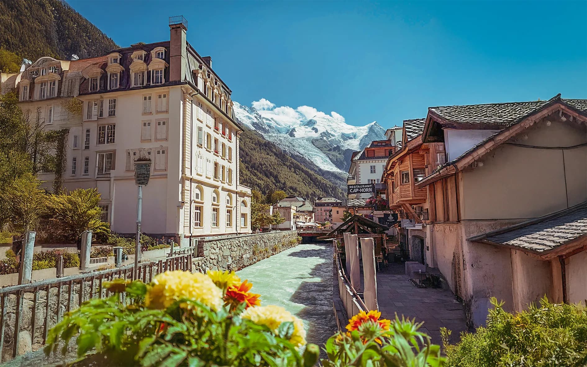 A picturesque Alpine village with buildings featuring both modern and traditional facades. A clear stream runs through the village, surrounded by greenery and colourful flowers. In the background, majestic mountains