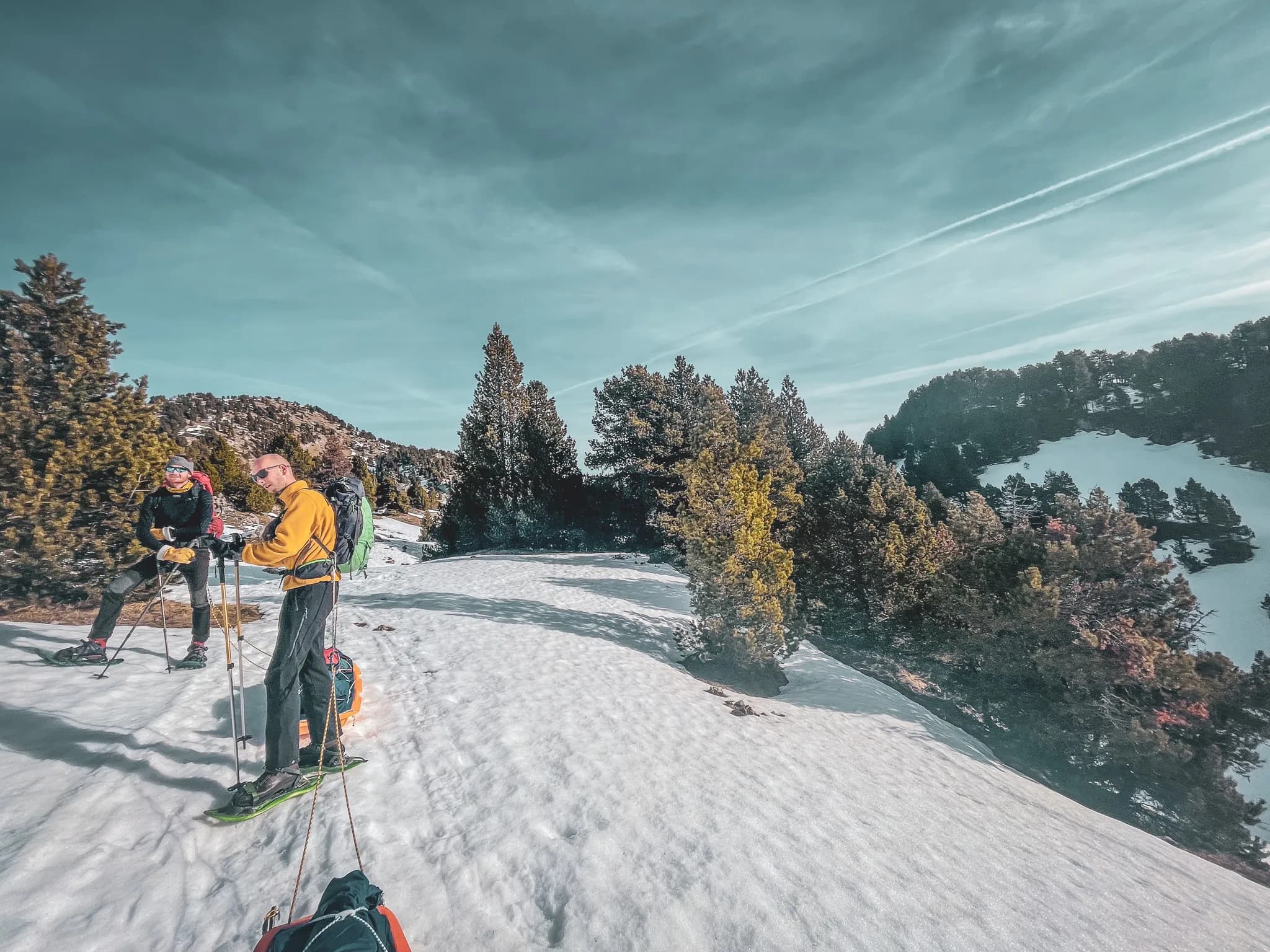 A snow-covered mountain landscape with two people in hiking gear. One is wearing a yellow jumper and carrying snowshoes, while the other is dressed in black with sunglasses. They are