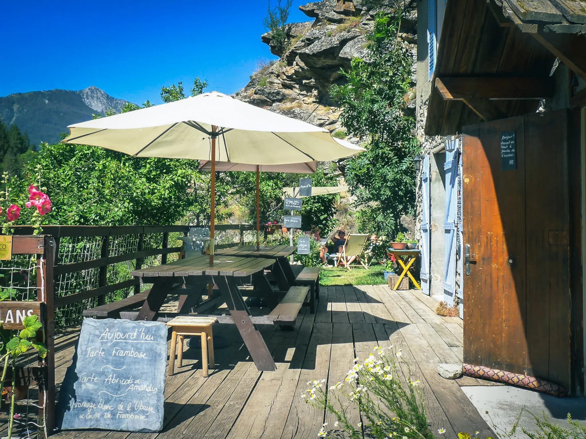 A relaxing outdoor area with a large wooden table under a white parasol, surrounded by greenery and flowers. To the left, a blackboard displays daily specialities such as pies. In the background