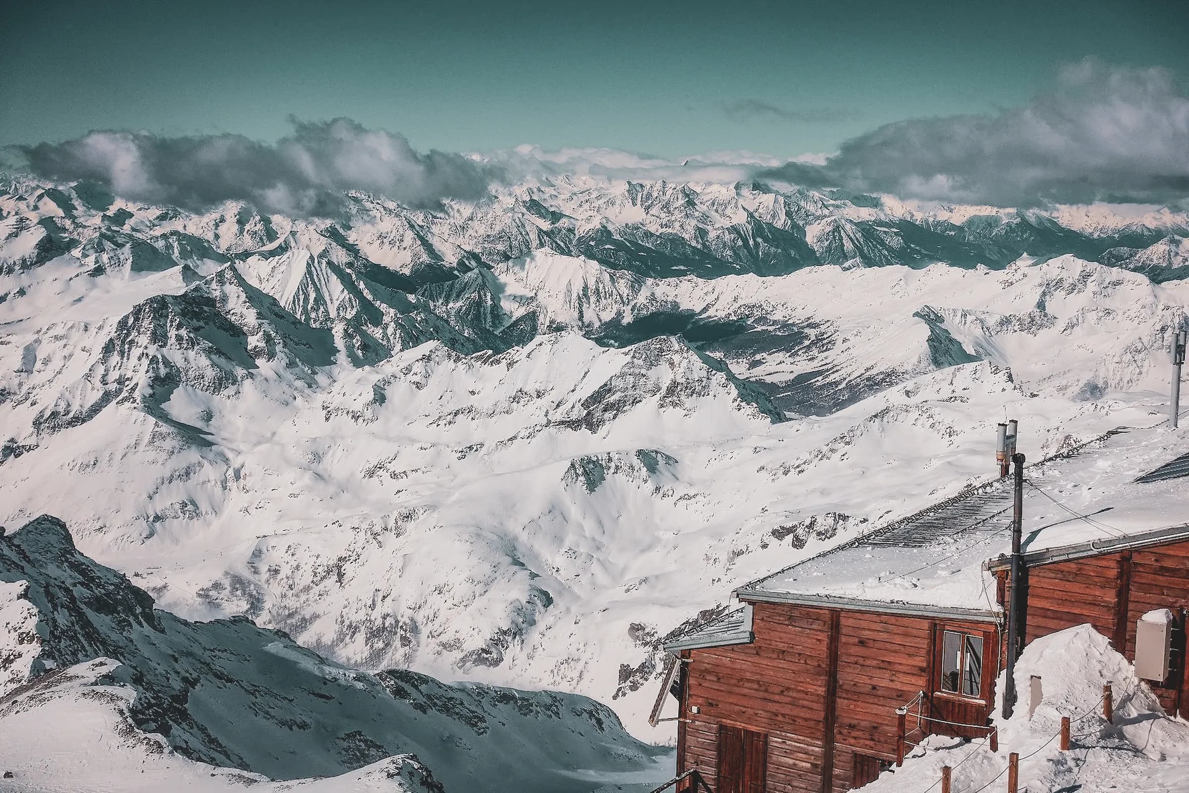A vast landscape of snow-capped mountains stretches as far as the eye can see, with majestic, craggy peaks covered in a thick layer of brilliant snow. The sky, tinged with shades of turquoise