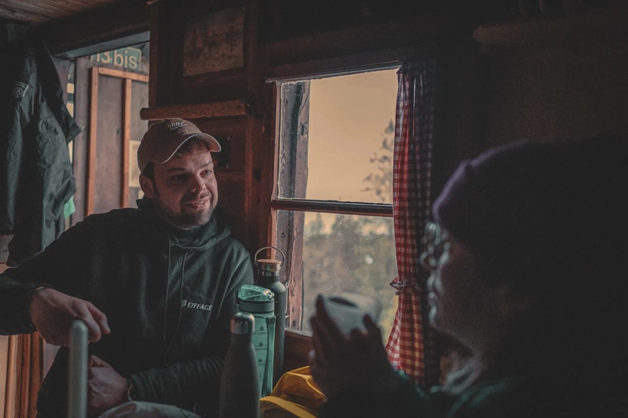 Deux personnes discutent chaleureusement à l'intérieur d'un chalet en bois, avec une vue sur la nature.