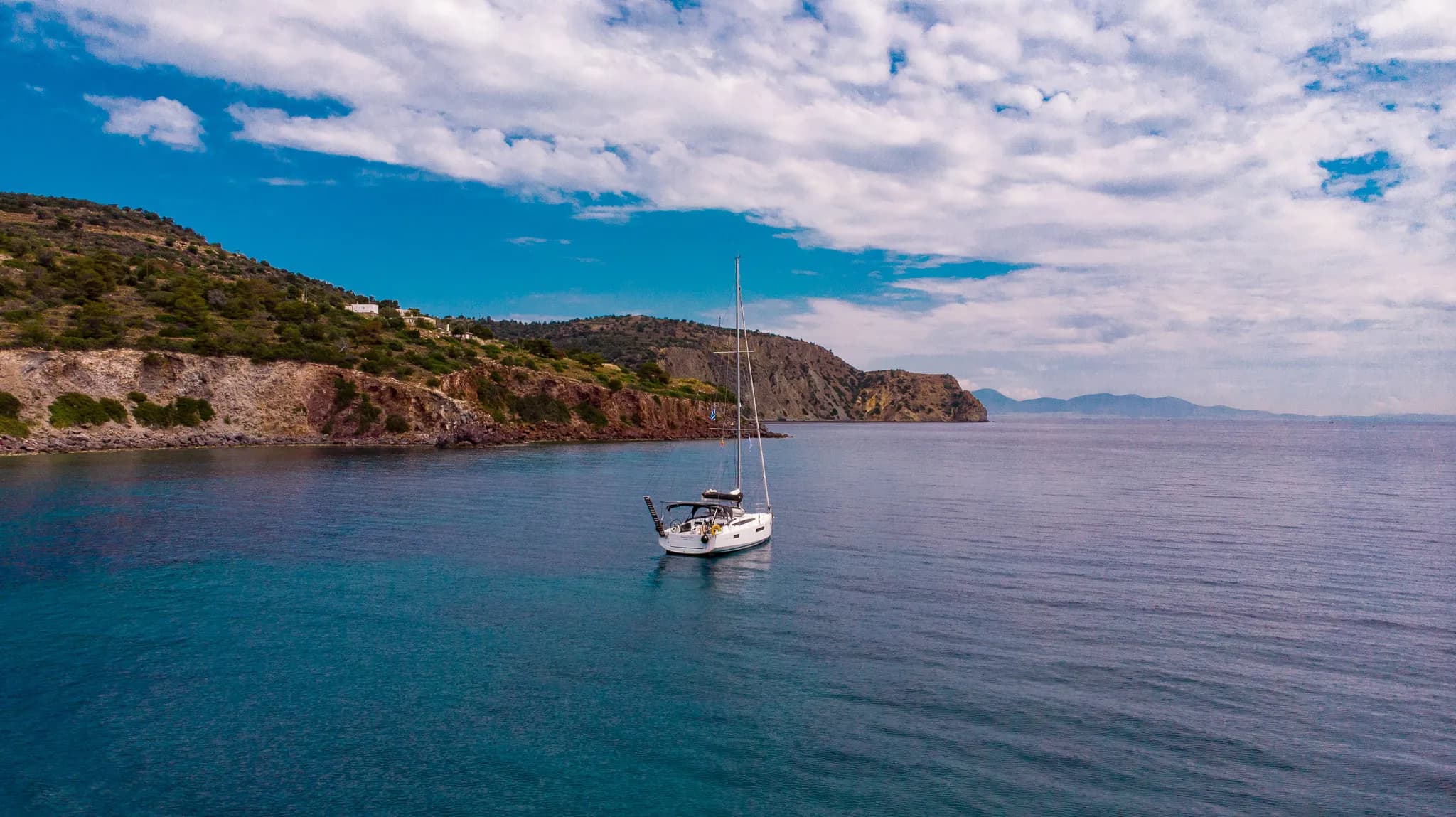 Voilier ancré dans une eau turquoise, bordé de collines verdoyantes sous un ciel bleu parsemé de nuages.
