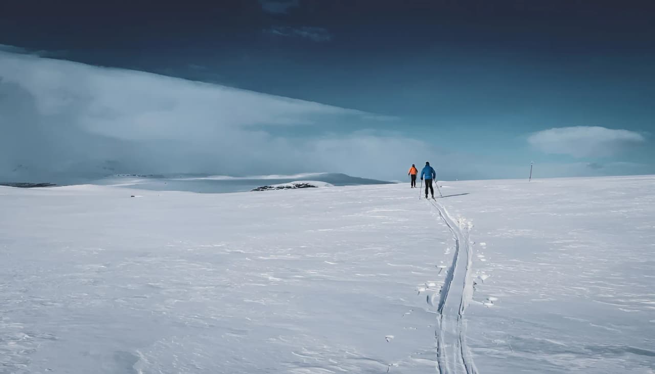 Twee skiërs doorkruisen een besneeuwd landschap onder een blauwe hemel in Kungsleden, Zweeds Lapland.
