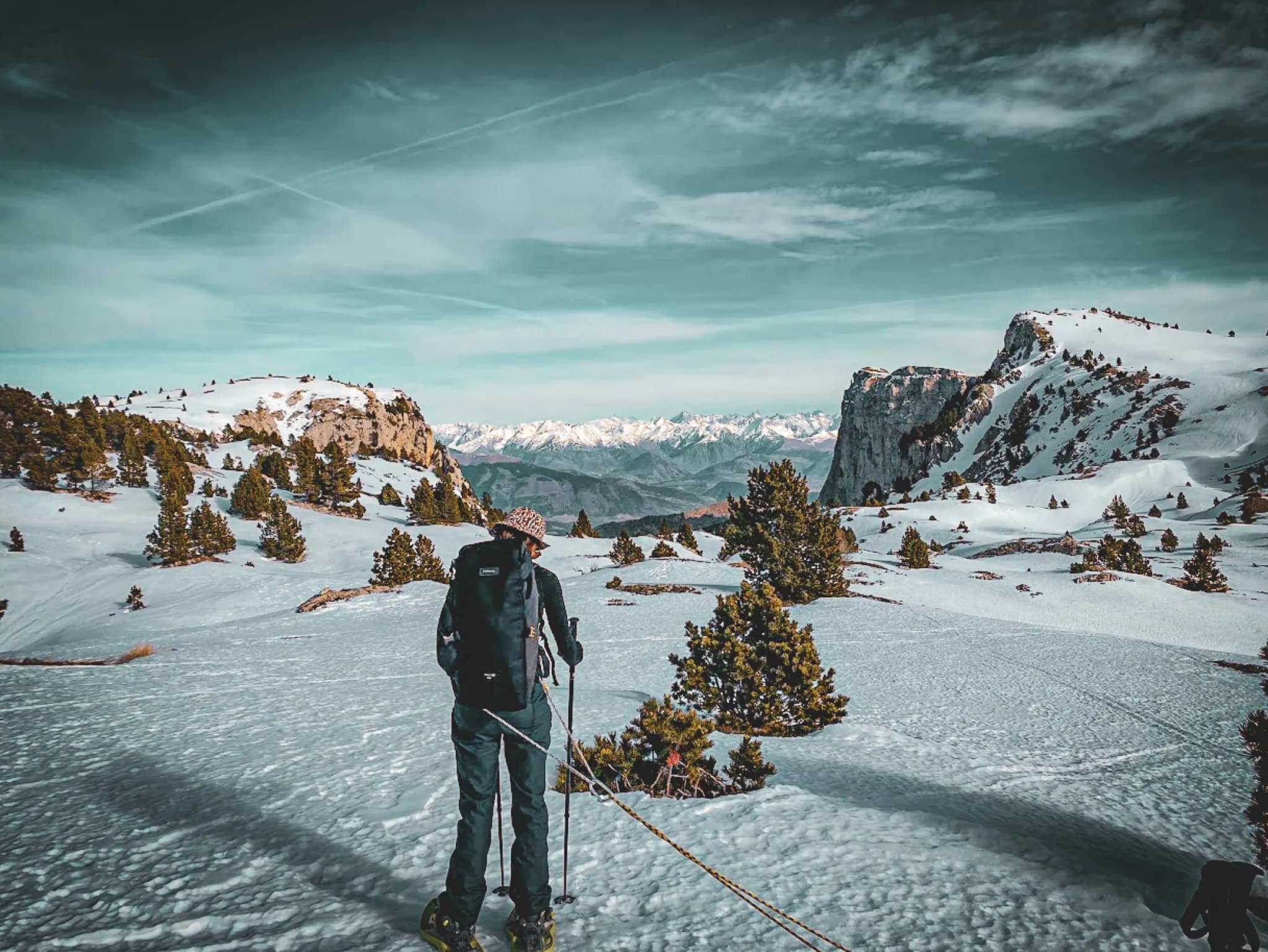 A man stands on snow-covered ground, looking out over a majestic Alpine landscape. Steep, snow-capped mountains loom on the horizon, while conifers dot the ground. L