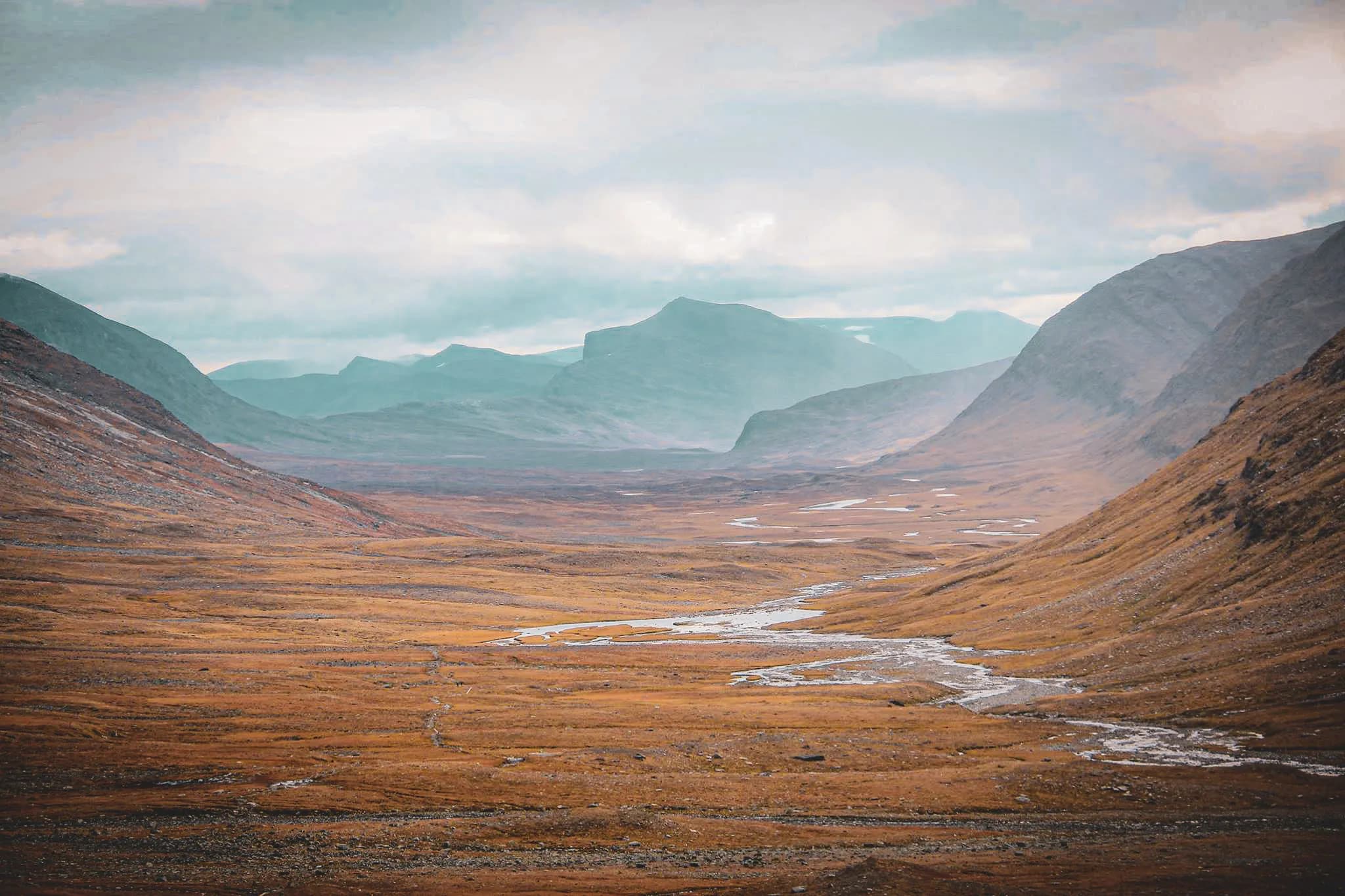 A vast, mountainous landscape, with wide valleys covered in golden grass and mountains in the background. A winding stream crosses the land, adding a touch of luminosity to the picture.