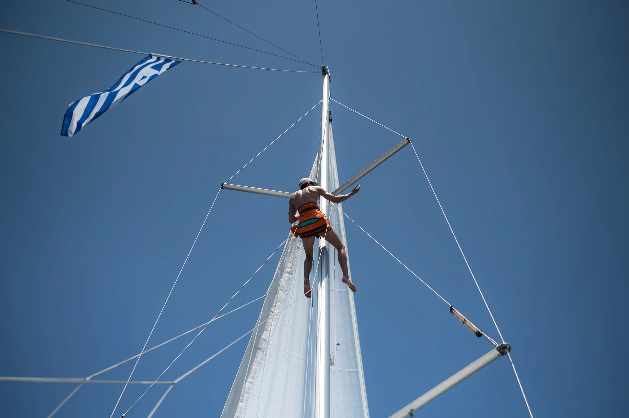 Un marin grimpe à la mât d'un voilier sous le ciel bleu de la Grèce, prêt pour l'aventure.