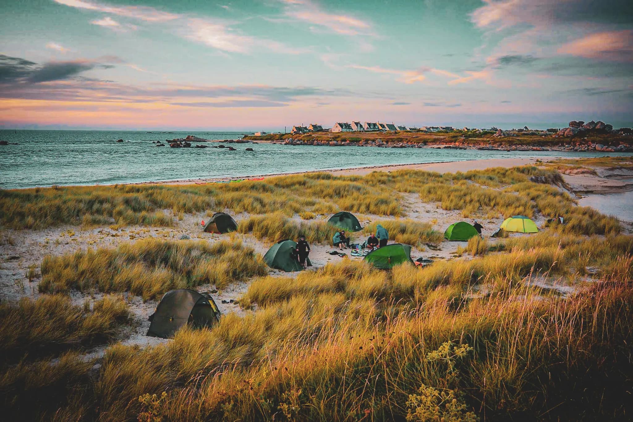 A campsite landscape by the sea, with several green tents arranged on a beach surrounded by tall grass. The ocean stretches to the horizon, under a sky of soft shades of blue and red.