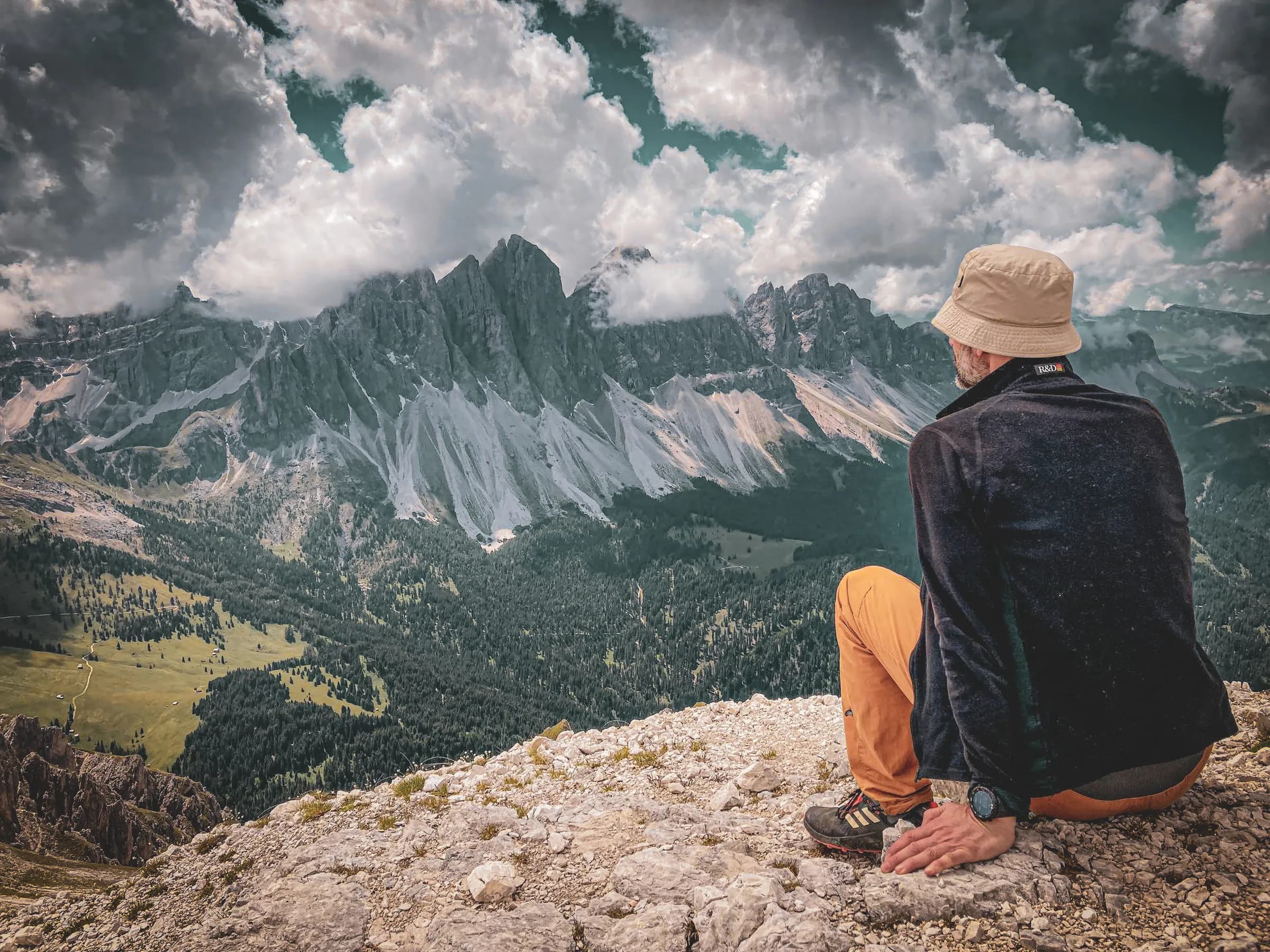 A man sitting on a rock overlooks an impressive mountain landscape, with craggy peaks and dramatic clouds. He is wearing a beige sun hat and dark jacket, as well as orange trousers.