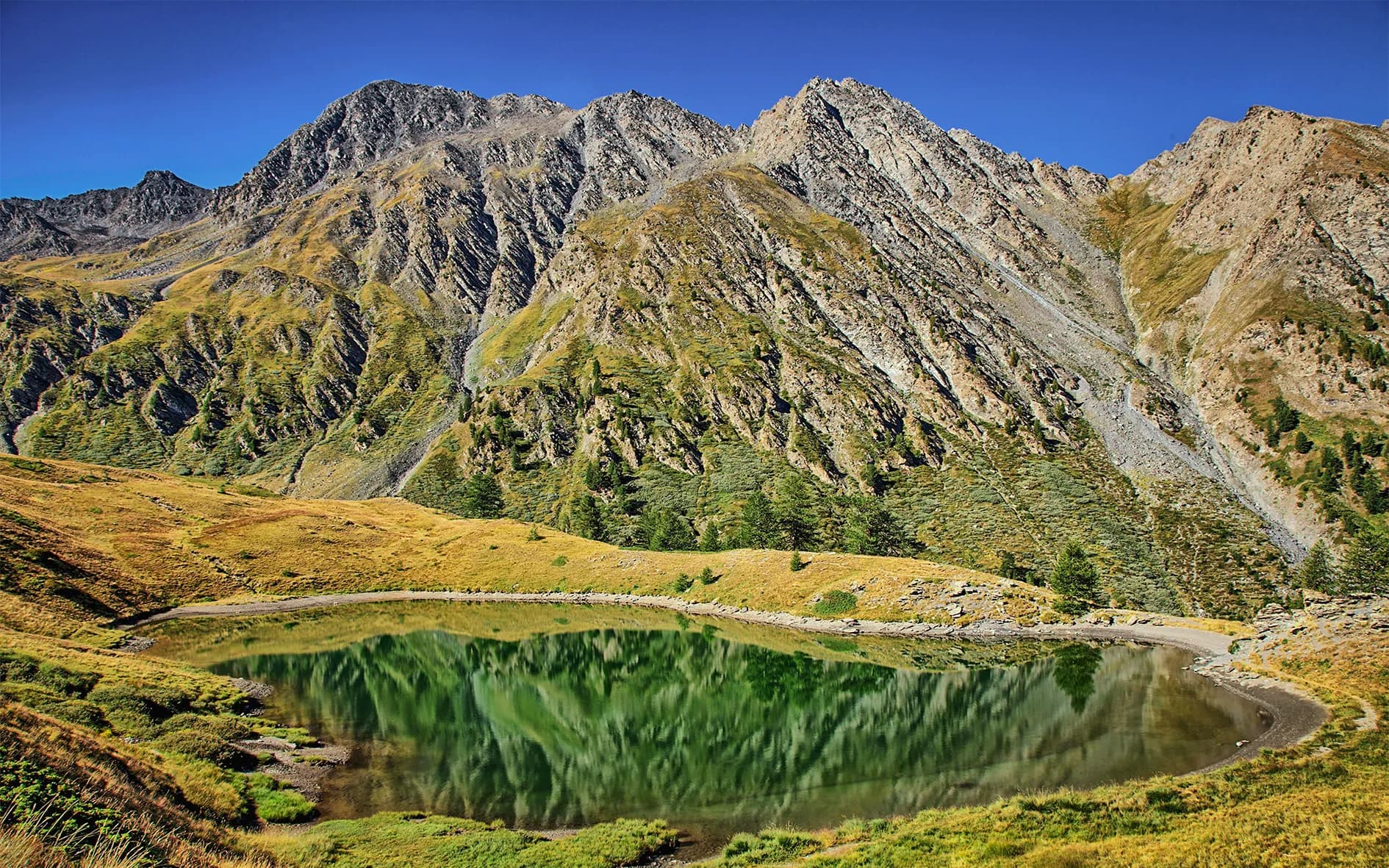 A majestic mountain landscape with a tranquil lake in the foreground. The emerald green waters of the lake reflect the surrounding rock faces, accentuated by a cloudless blue sky. The mountains