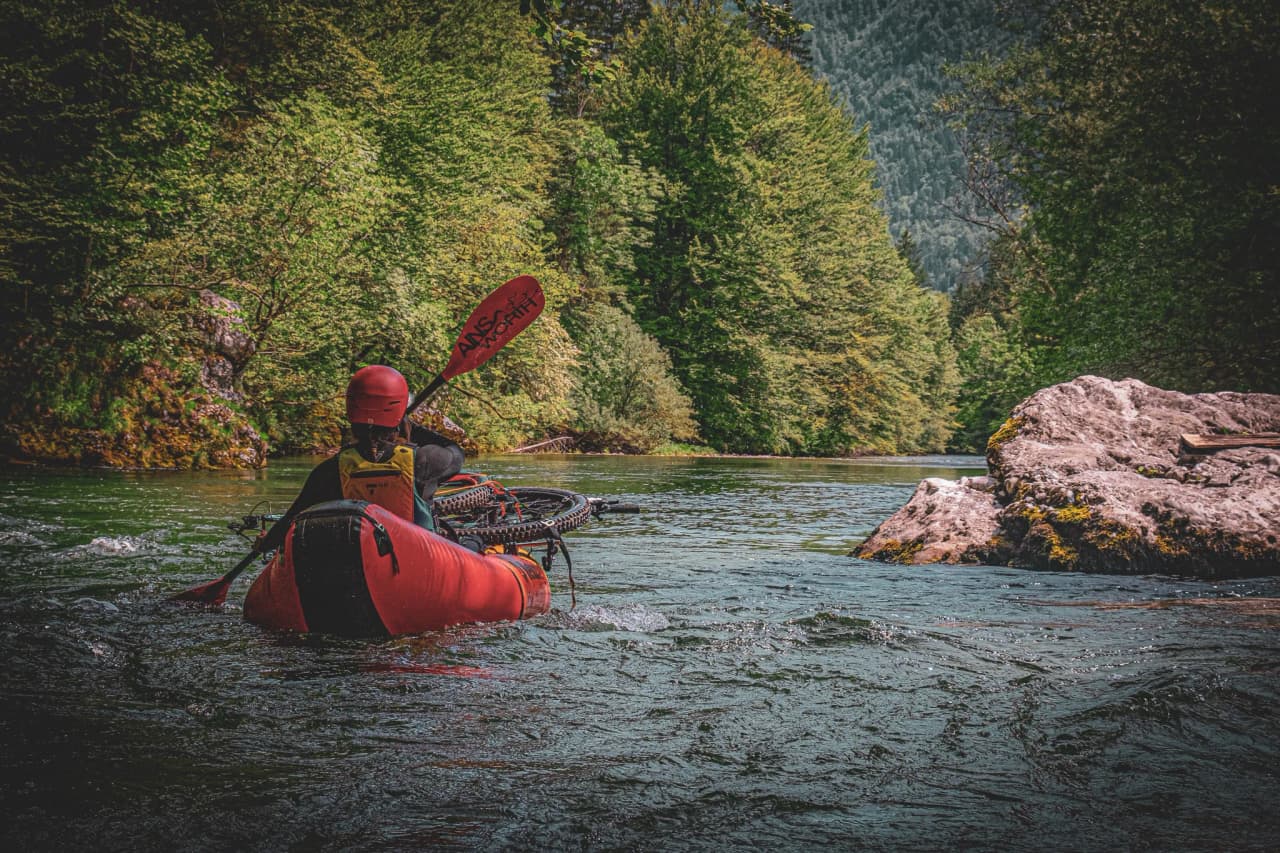 A red canoe moves gently along a river surrounded by lush greenery. One person, wearing a red helmet and holding a paddle, sits at the back of the canoe, while the other, wearing a red helmet and holding a paddle, sits at the back of the canoe.