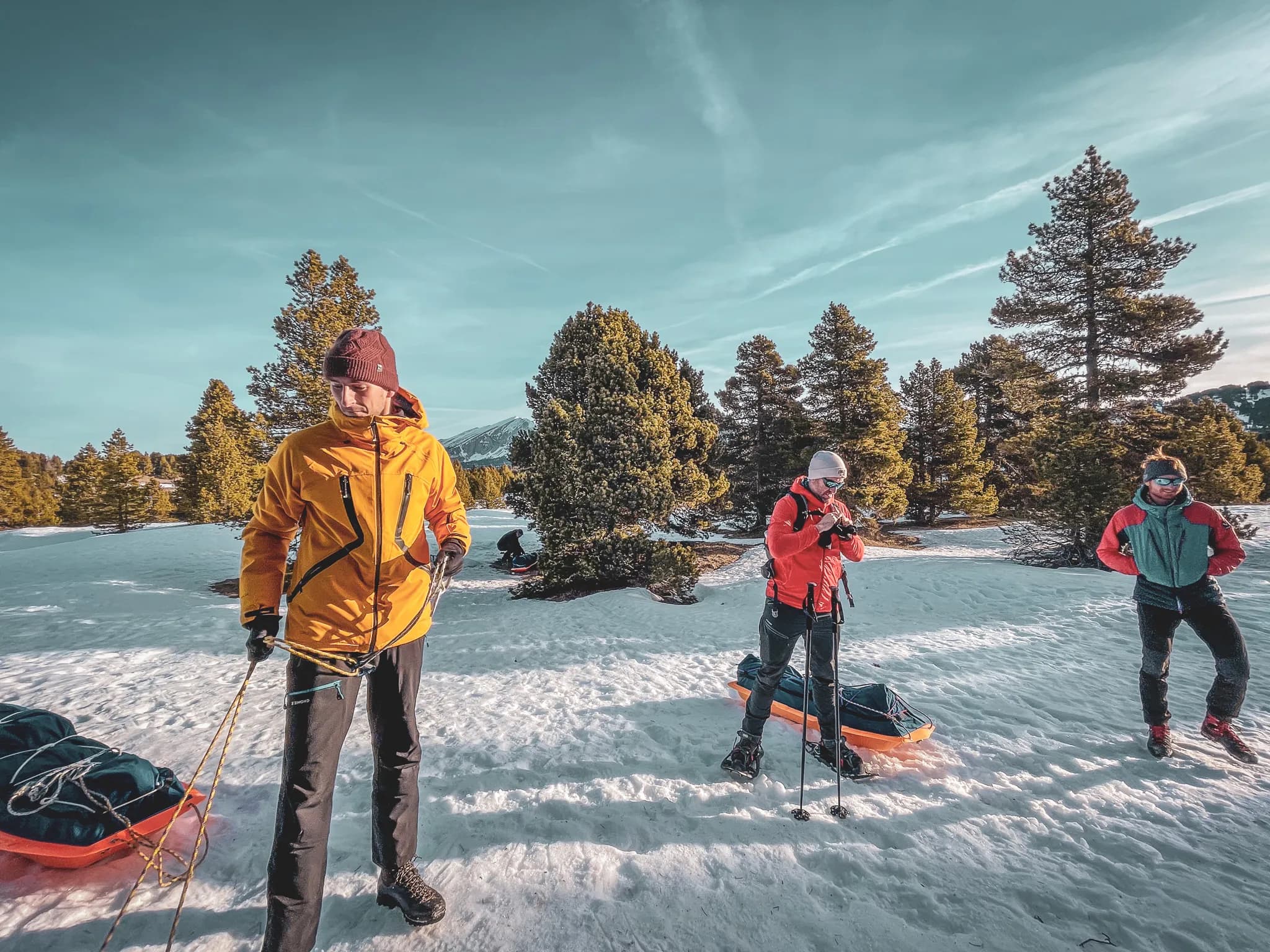 A group of people in full activity on snow, wearing appropriate outdoor clothing. One of the members is wearing a yellow jacket and holding ropes, while another is wearing a red jacket and looking at his camera. A