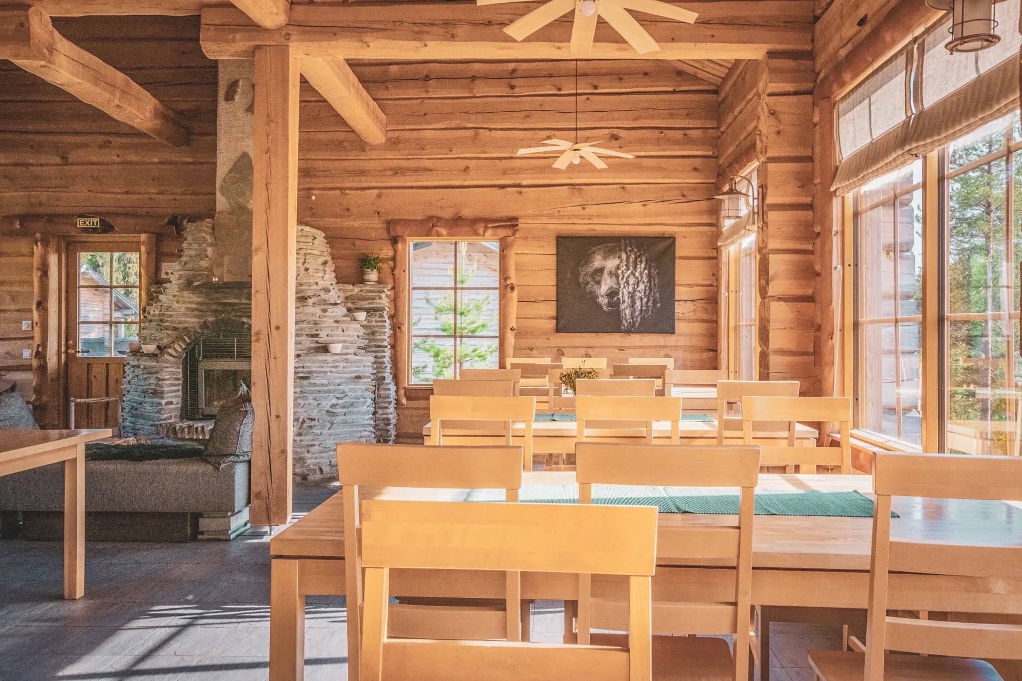 A warm wooden interior with log walls and a dark wood floor. In the centre, several light wood tables are arranged with matching chairs. In the background, a large stone stove is the centrepiece.