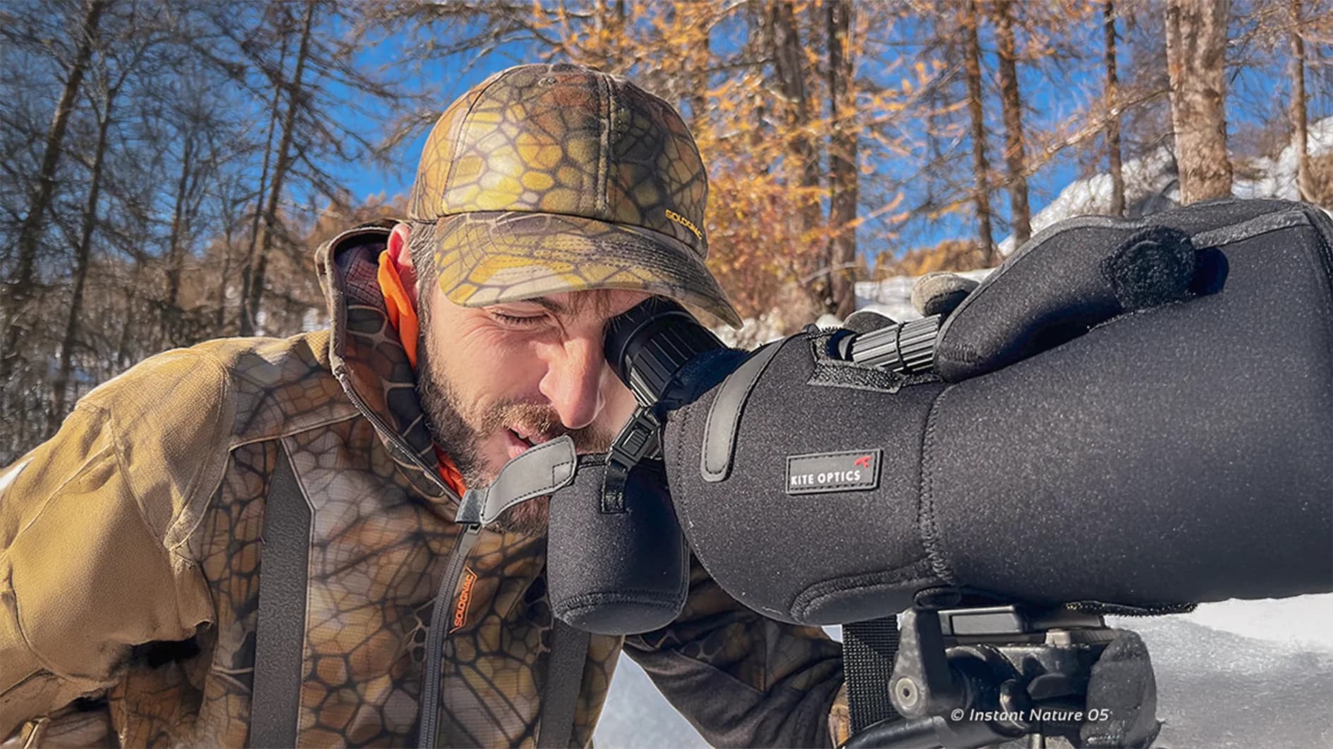 A man in camouflage is observing through a telescope on a tripod in a snowy environment. He is wearing a patterned cap in shades of brown and green, and his expression shows concentration. At