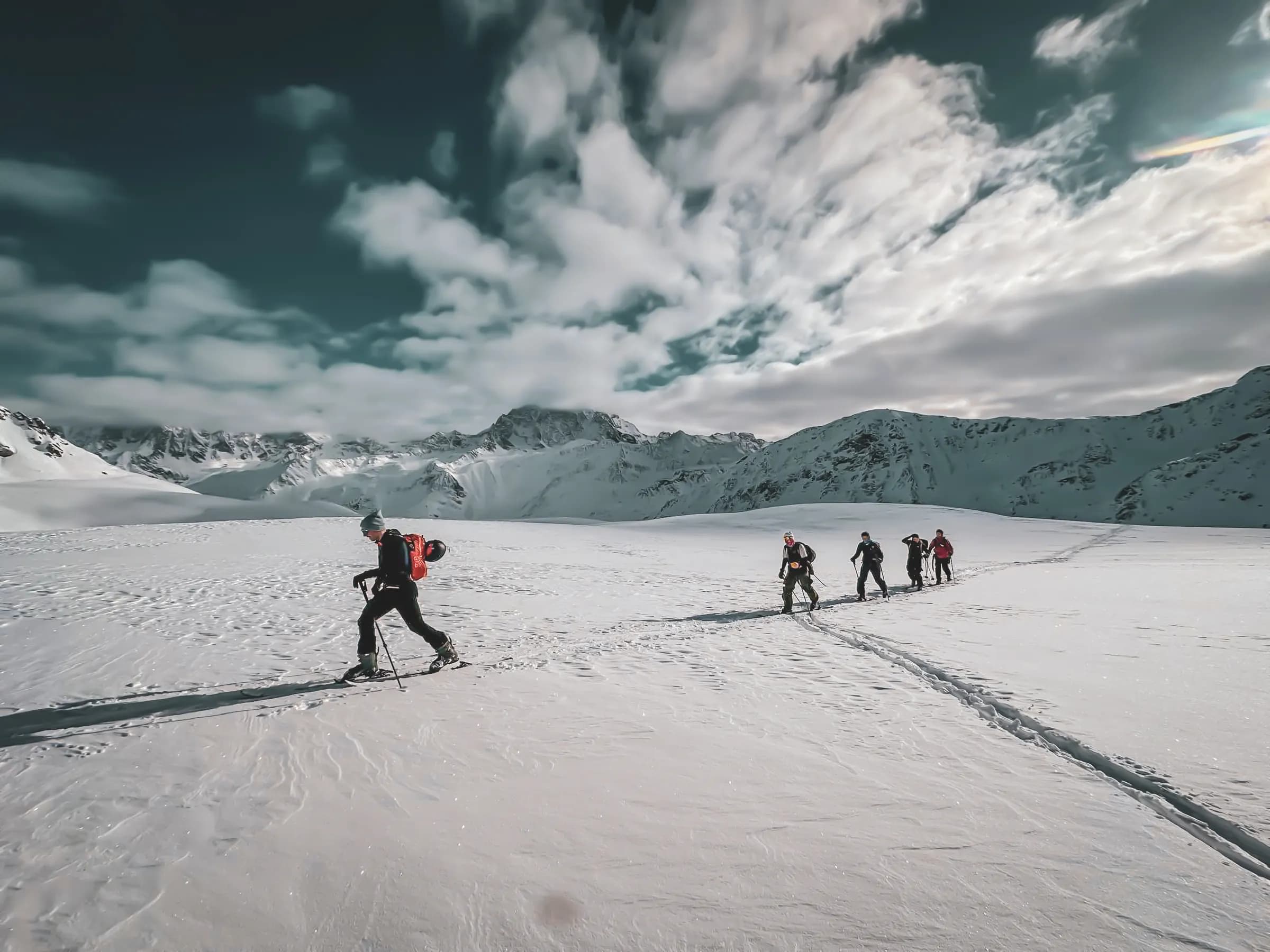A group of hikers are walking across a vast expanse of snow, surrounded by majestic mountains under a cloudy sky. One of the hikers, at the front, is carrying a red rucksack and