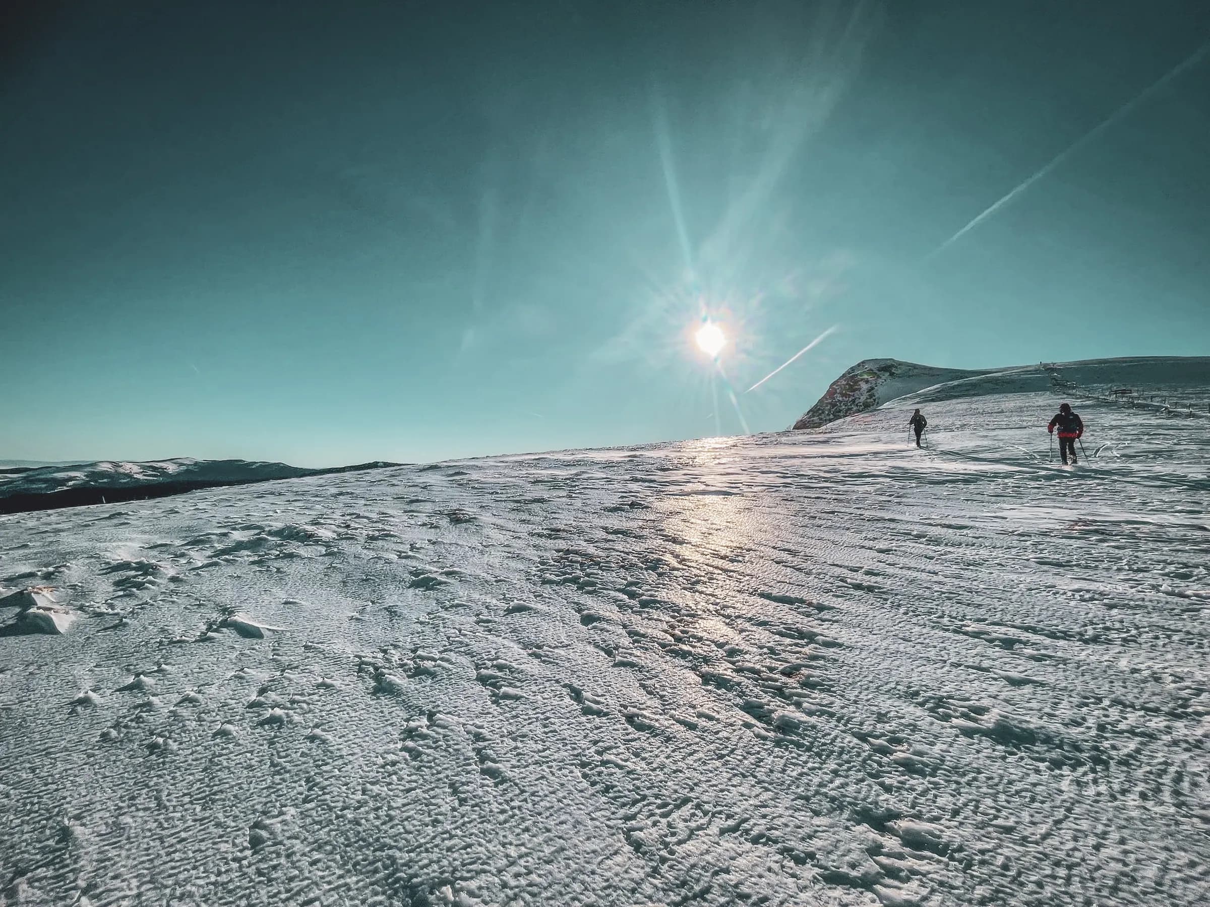Een besneeuwd landschap met een heldere hemel, waar twee wandelaars zich een weg banen over een besneeuwd oppervlak. De zon schijnt fel, waardoor reflecties op de sneeuw ontstaan. Bergen in de verte komen in zicht