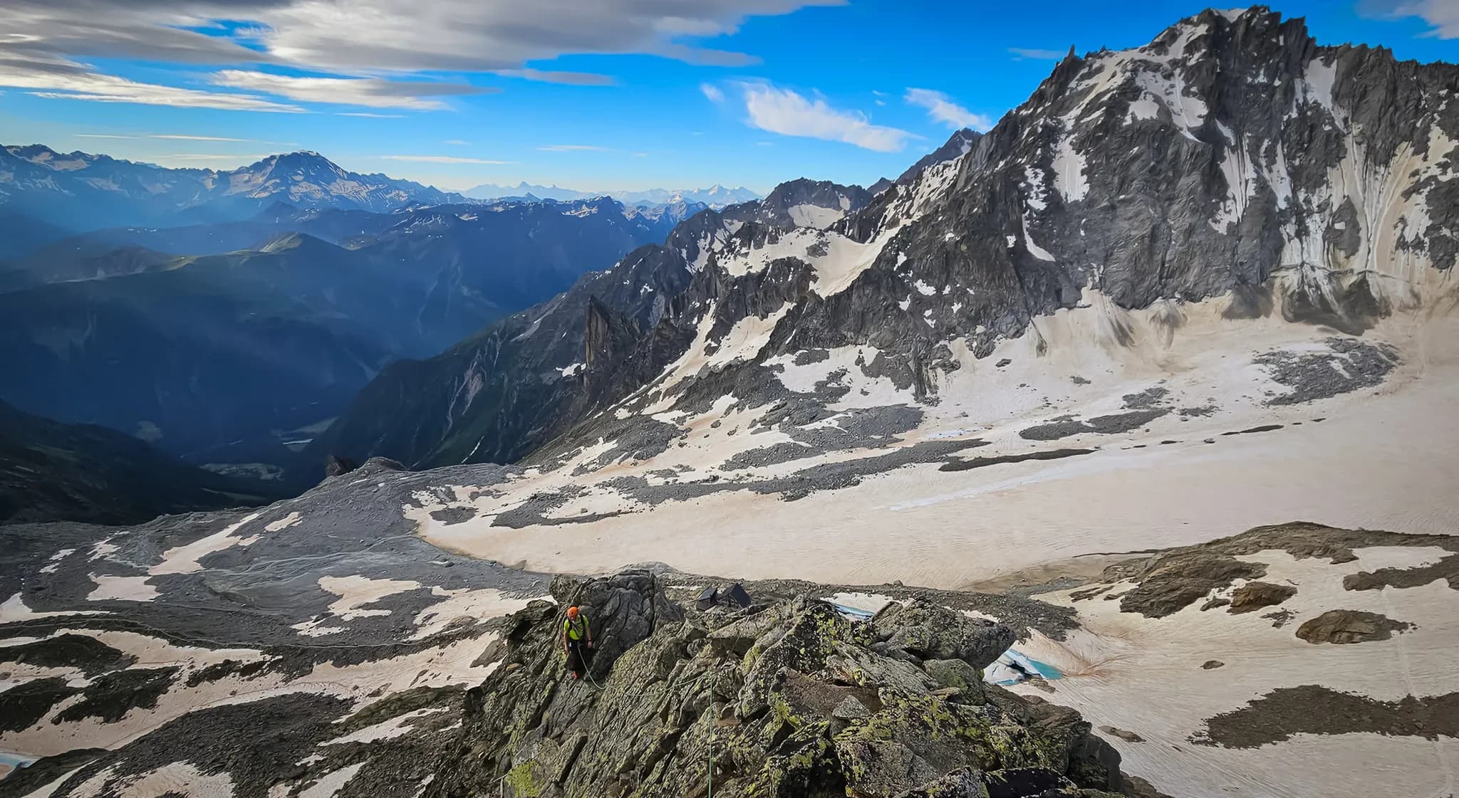 A spectacular mountain landscape with steep peaks and glaciers. In the foreground, a well-equipped climber in orange and yellow clothing scrambles over rocky outcrops, while in the background, valleys and glaciers are visible.
