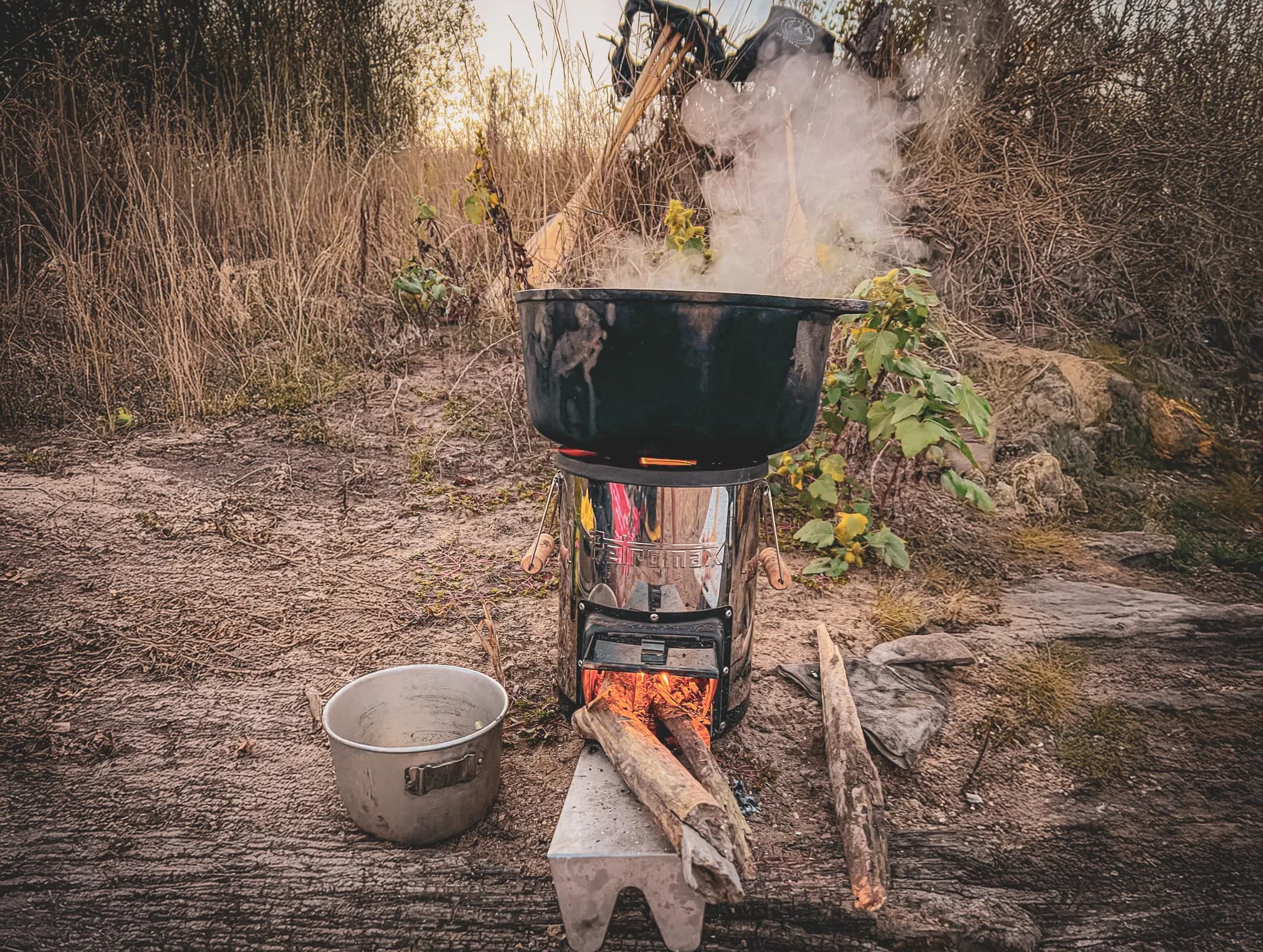 A metal camping stove is set up on natural ground, surrounded by vegetation. A large black saucepan sits on the stove, giving off steam. Underneath, pieces of wood are used