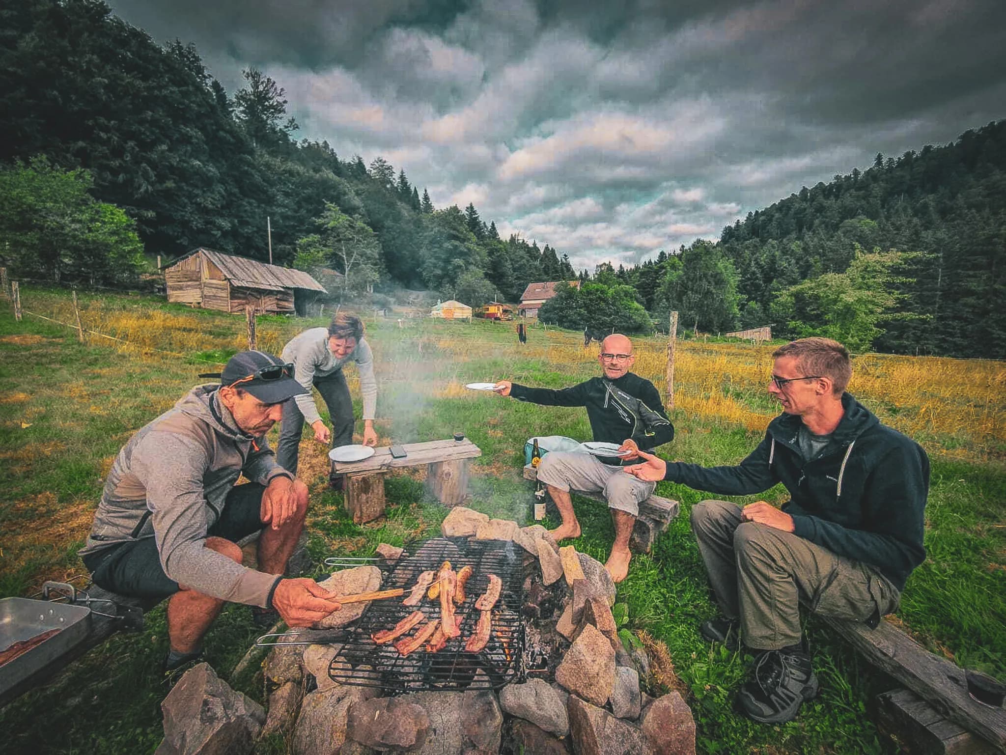 A group of four people gather around a campfire in the middle of nowhere. Two of them are sitting on benches, while two others are standing by the barbecue, cooking slices of