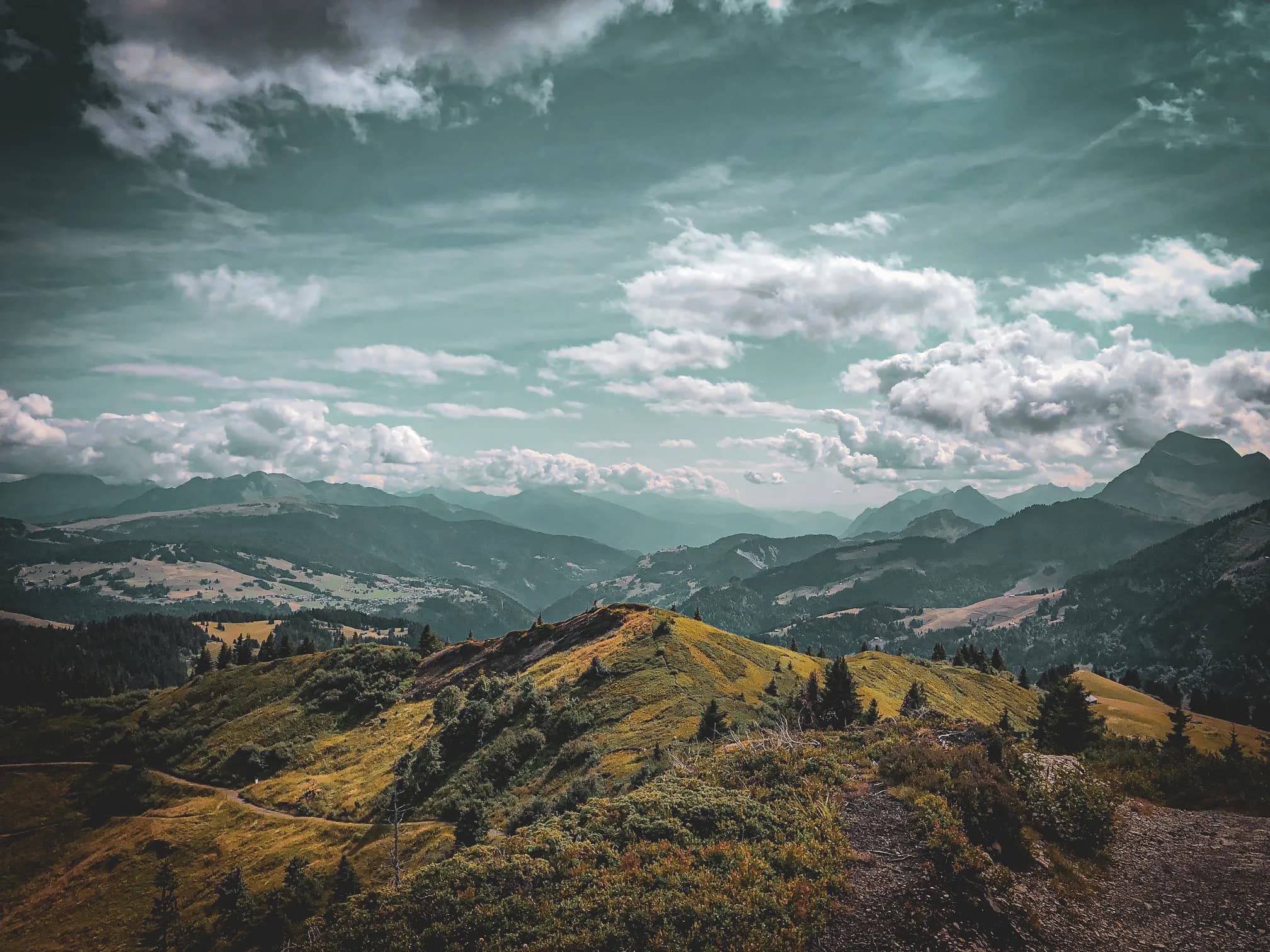 A vast mountain landscape with green hills and scattered forests. In the distance, mountain ranges with slightly misty summits can be seen under a cloudy sky. The clouds