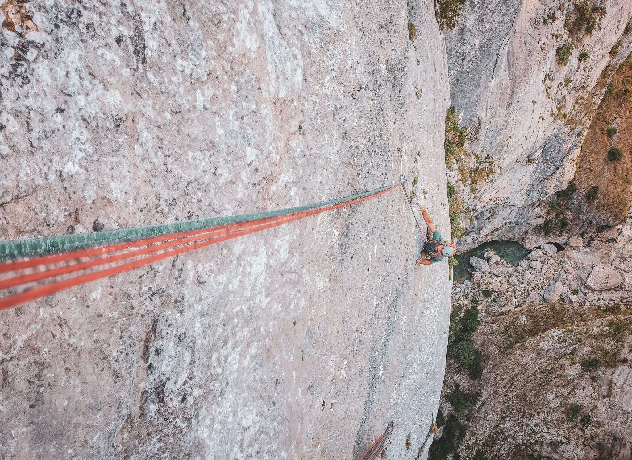 A steep cliff with a climber in full effort, secured by red and green ropes. The climber is ascending, clinging to the rock face, while the landscape in