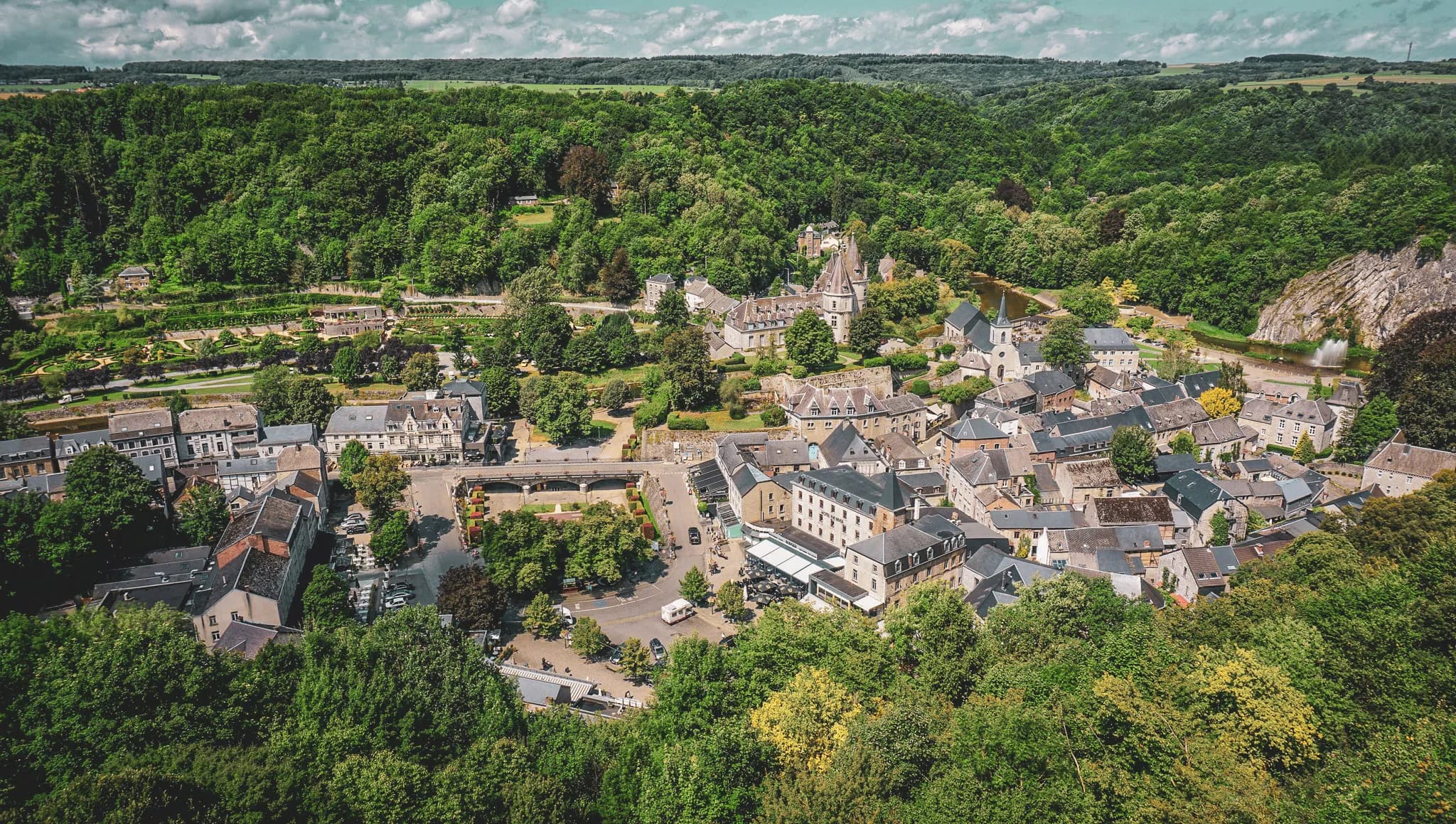 A picturesque village surrounded by green hills, with slate-roofed houses and historic buildings. There is a small river winding through the landscape, and well-landscaped green spaces,