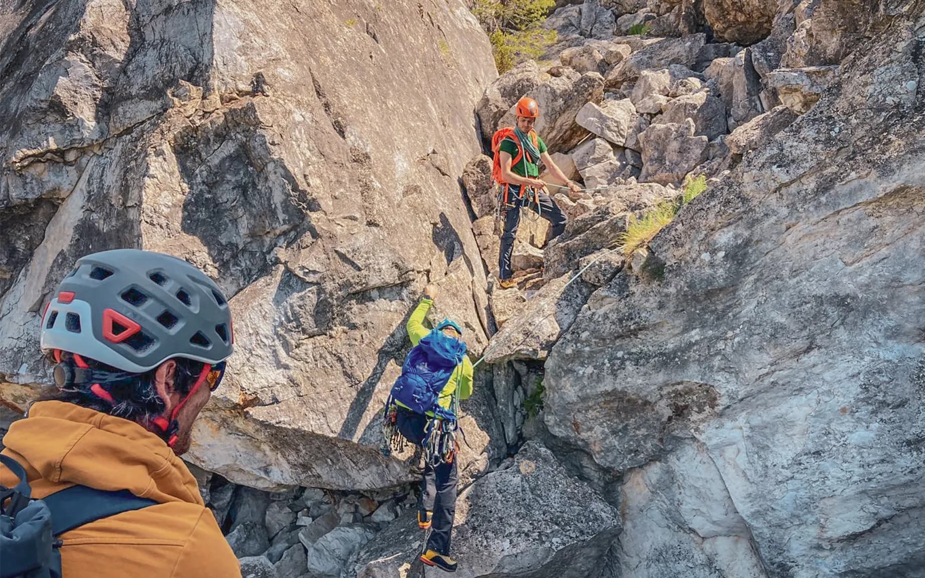 A group of three climbers are on a steep rock face in the middle of nowhere. Two of them are in action; one appears to be climbing a difficult section, while the other, wearing an orange helmet, is securing his position.