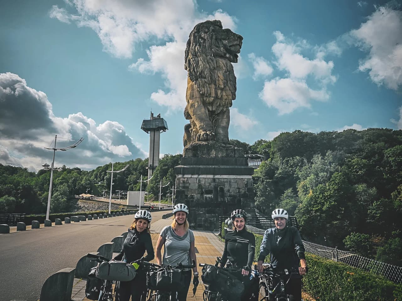 Quatre cyclistes souriants avec leurs vélos, devant une grande statue de lion, sous un ciel ensoleillé.