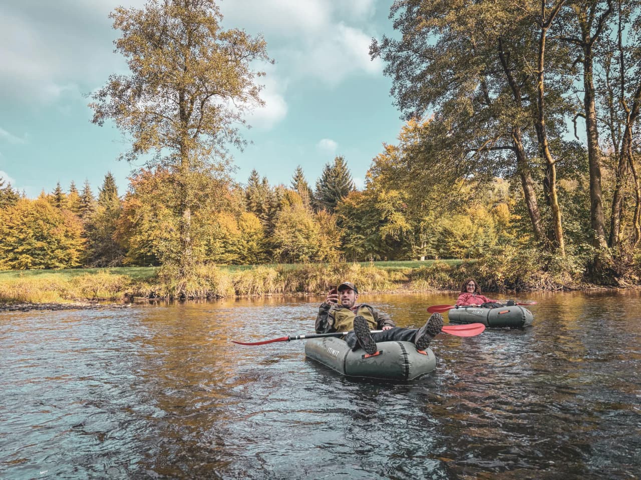 Two people relax in a packraft on a river surrounded by autumn-coloured forests.