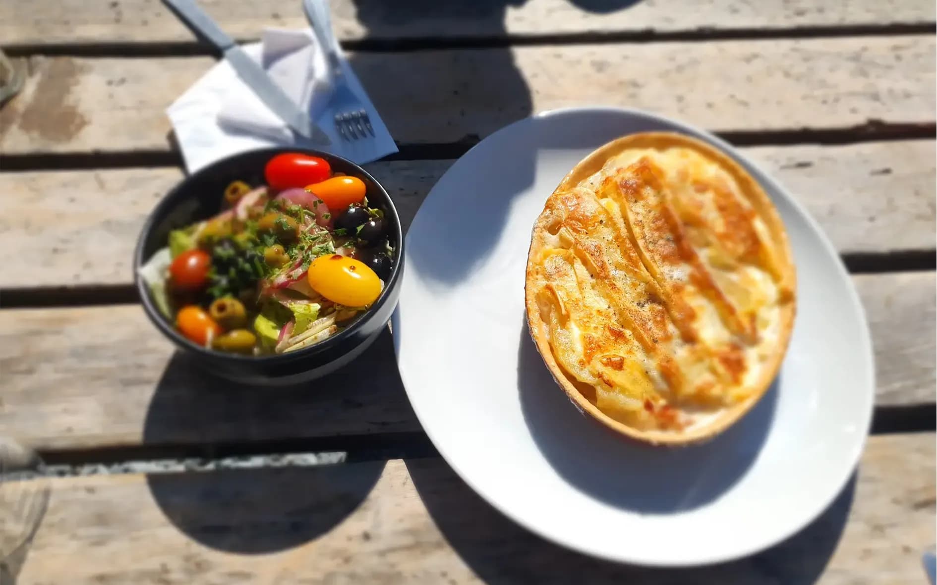 An appetising dish of crustless potato pie, golden brown and slightly crispy on top, is served on a white plate. Next to it, a black bowl holds a colourful salad topped with