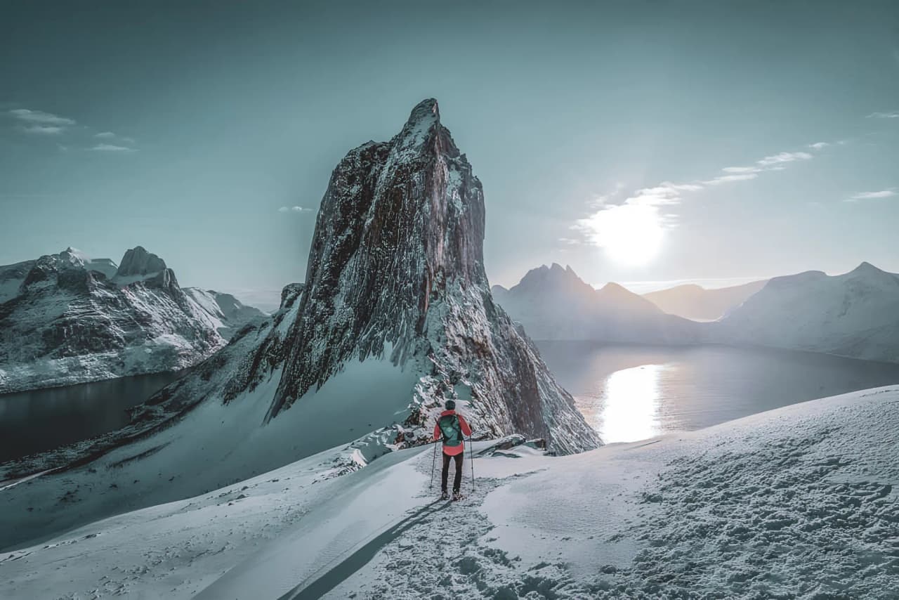 A snowshoe hiker facing an icy fjord under a bright dawn sky in Senja.