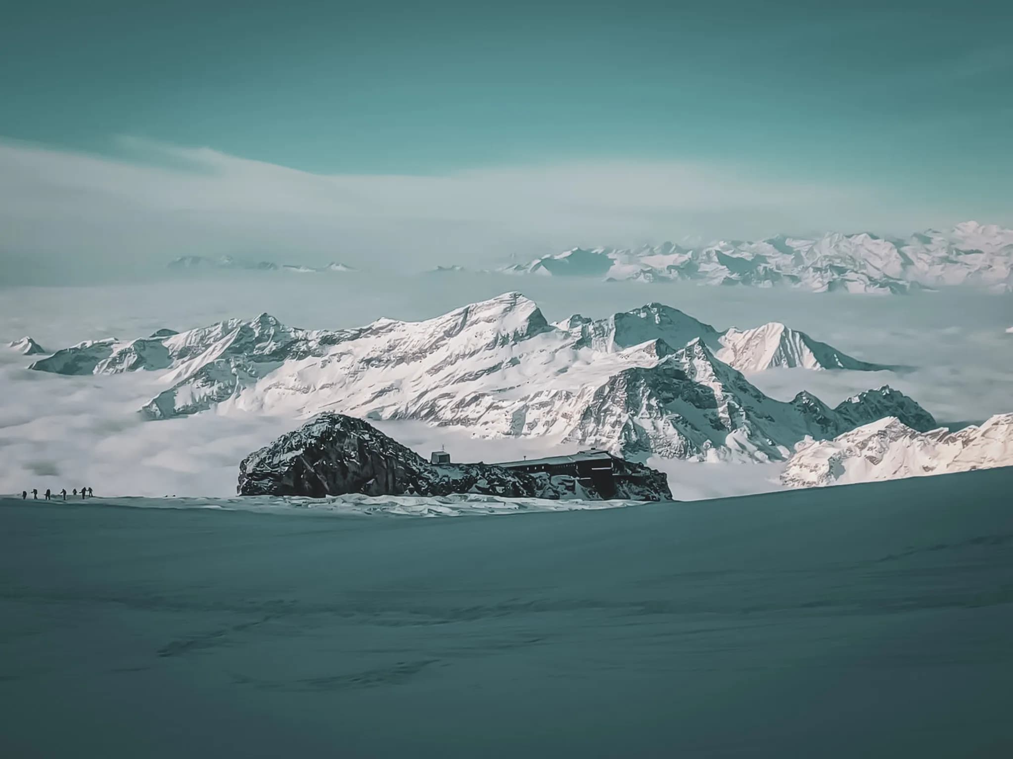 Een majestueus Alpenlandschap met besneeuwde bergen die boven een zee van wolken uitsteken. Op de voorgrond is een imposante rots te zien met daarop een gebouw. Aan de horizon menselijke silhouetten