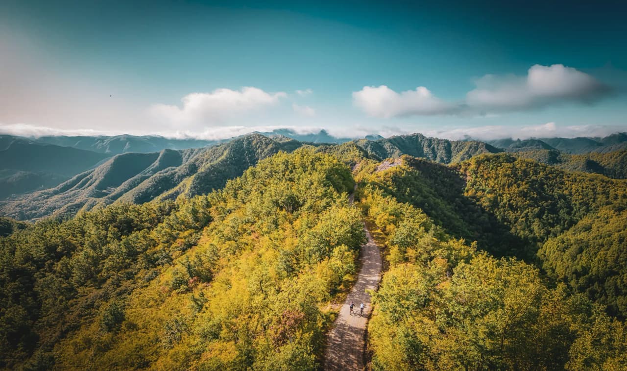 A majestic mountain landscape, with green hills and dense trees, unfolds beneath a clear blue sky dotted with a few clouds.