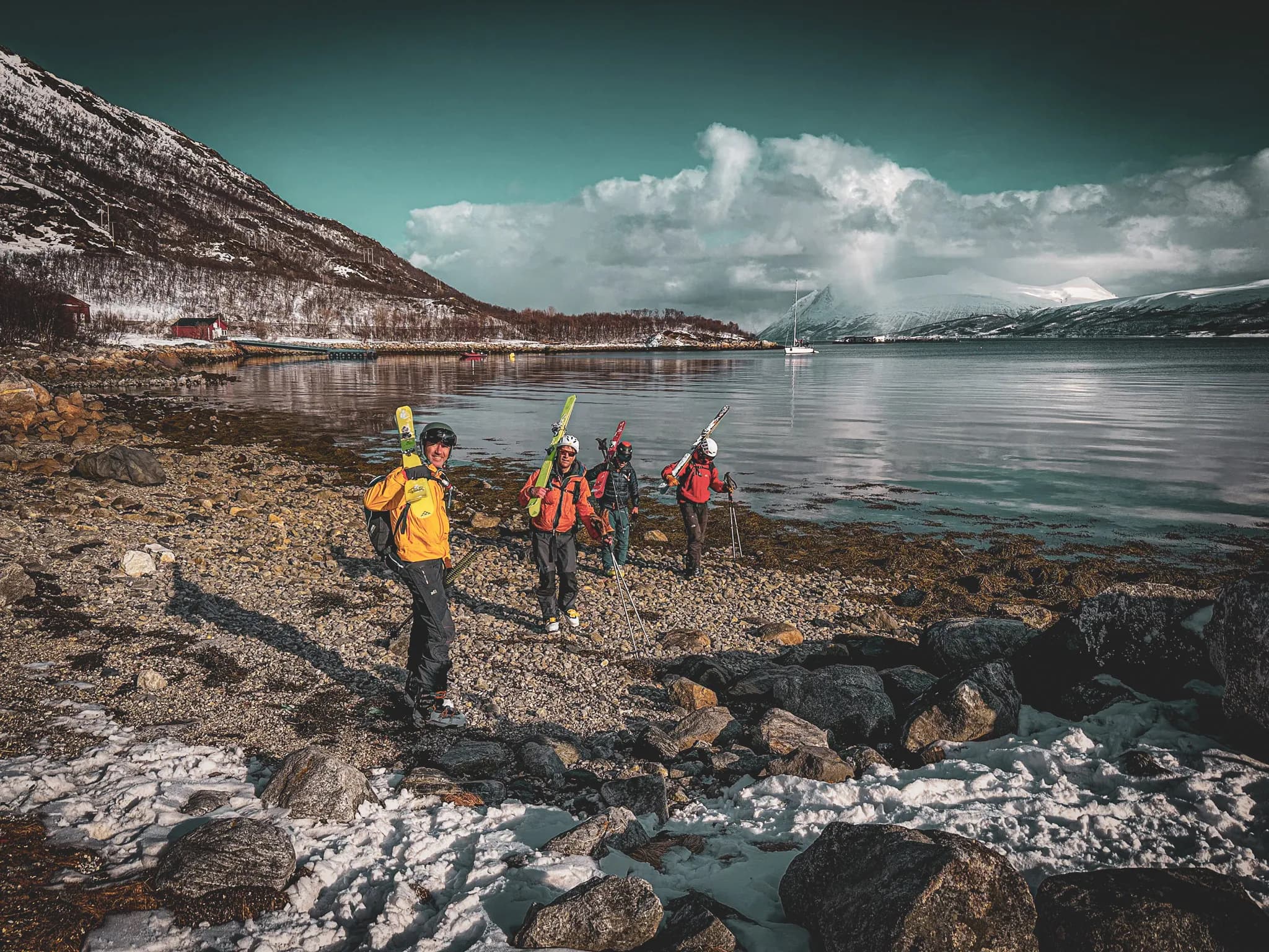 Un groupe de skieurs marche sur une plage de galets, portant leurs skis et équipements. Ils sont vêtus de vêtements d'extérieur colorés, avec des vestes rouges et jaunes. En arrière-plan, des montagnes enneigées s'élè