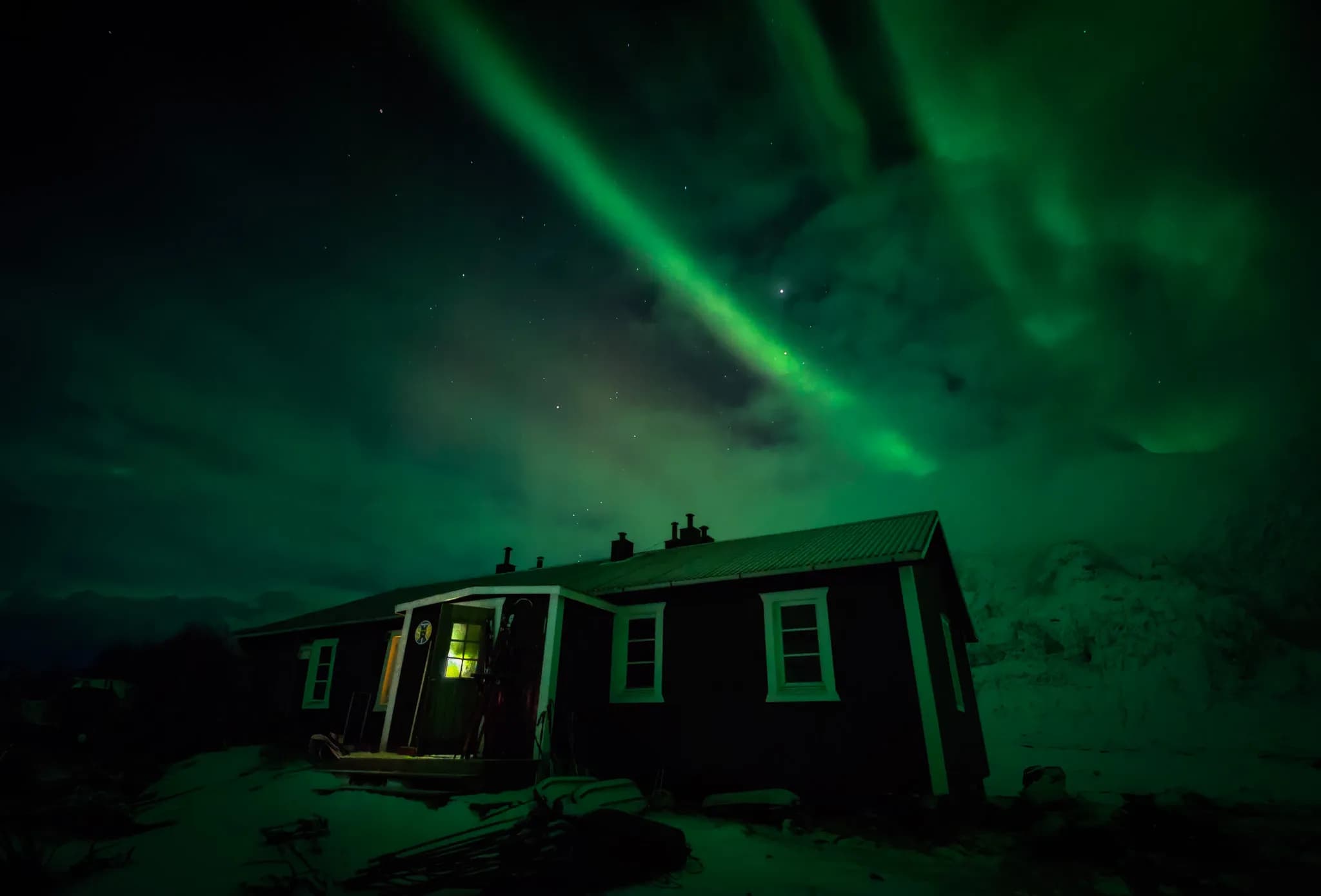 An STF wooden chalet stands in a snowy landscape, under a night sky illuminated by the northern lights in shades of green. Yellow light from a window illuminates the entrance.