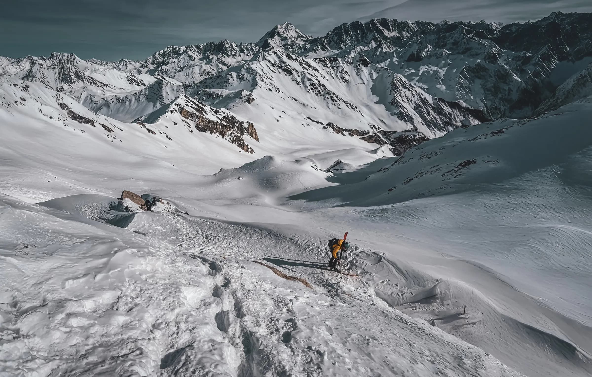 A vast mountain landscape covered in snow, with gentle slopes and steep peaks. In the foreground, you can see a skier in a yellow outfit, making his way up a slope. In the background