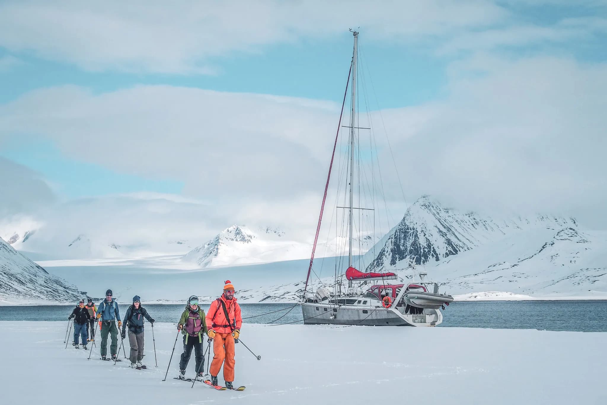 Un groupe de skieurs s'avance sur un paysage enneigé, tirant des traîneaux derrière eux. Les membres du groupe portent des vêtements colorés et variés, adaptés aux conditions froides. À l'arrière-plan, un vo