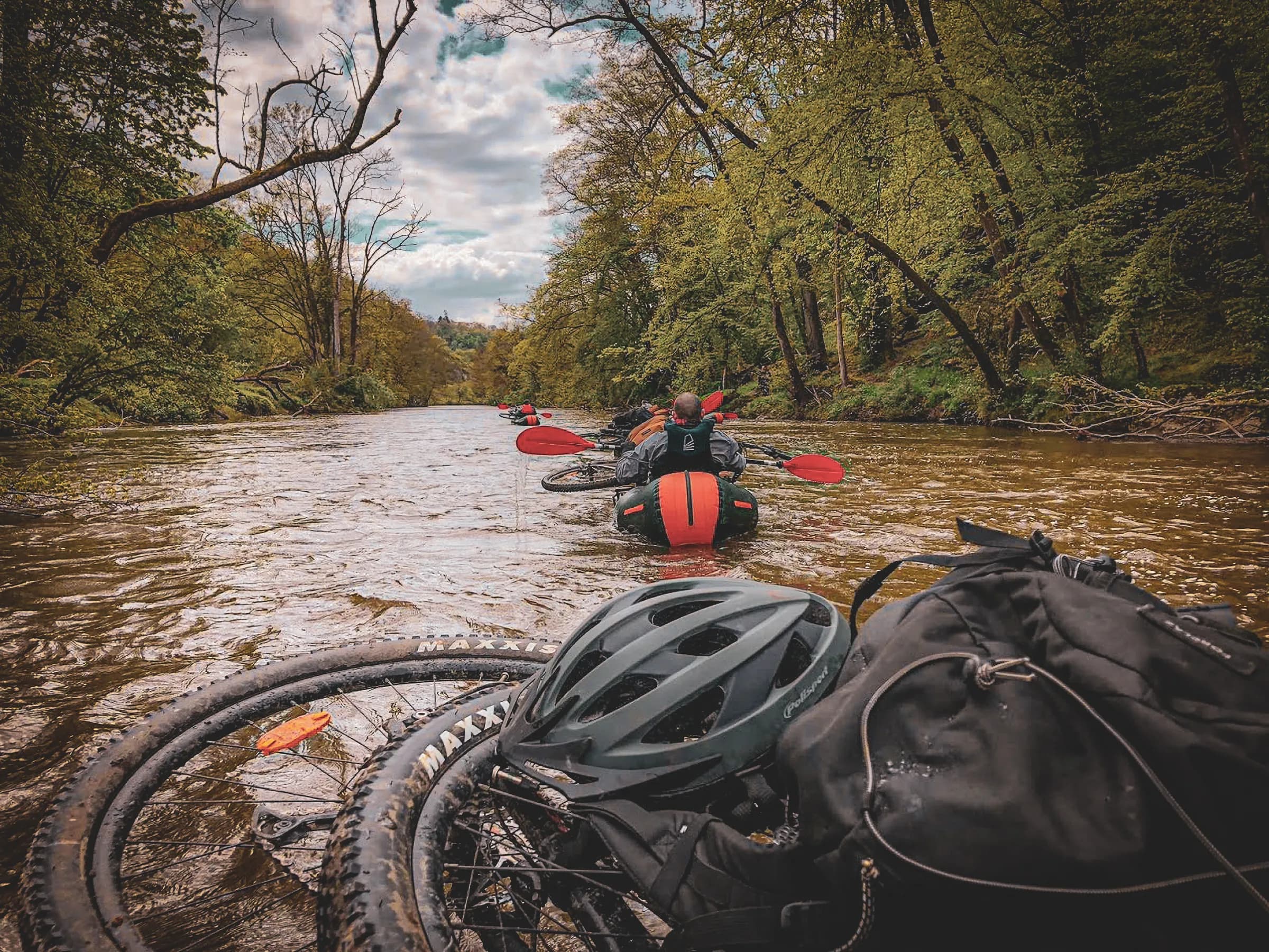 A group of kayakers are paddling on a river surrounded by greenery, while at the front of the image are bicycle tyres and a helmet. The water is slightly choppy, reflecting the cloudy sky that