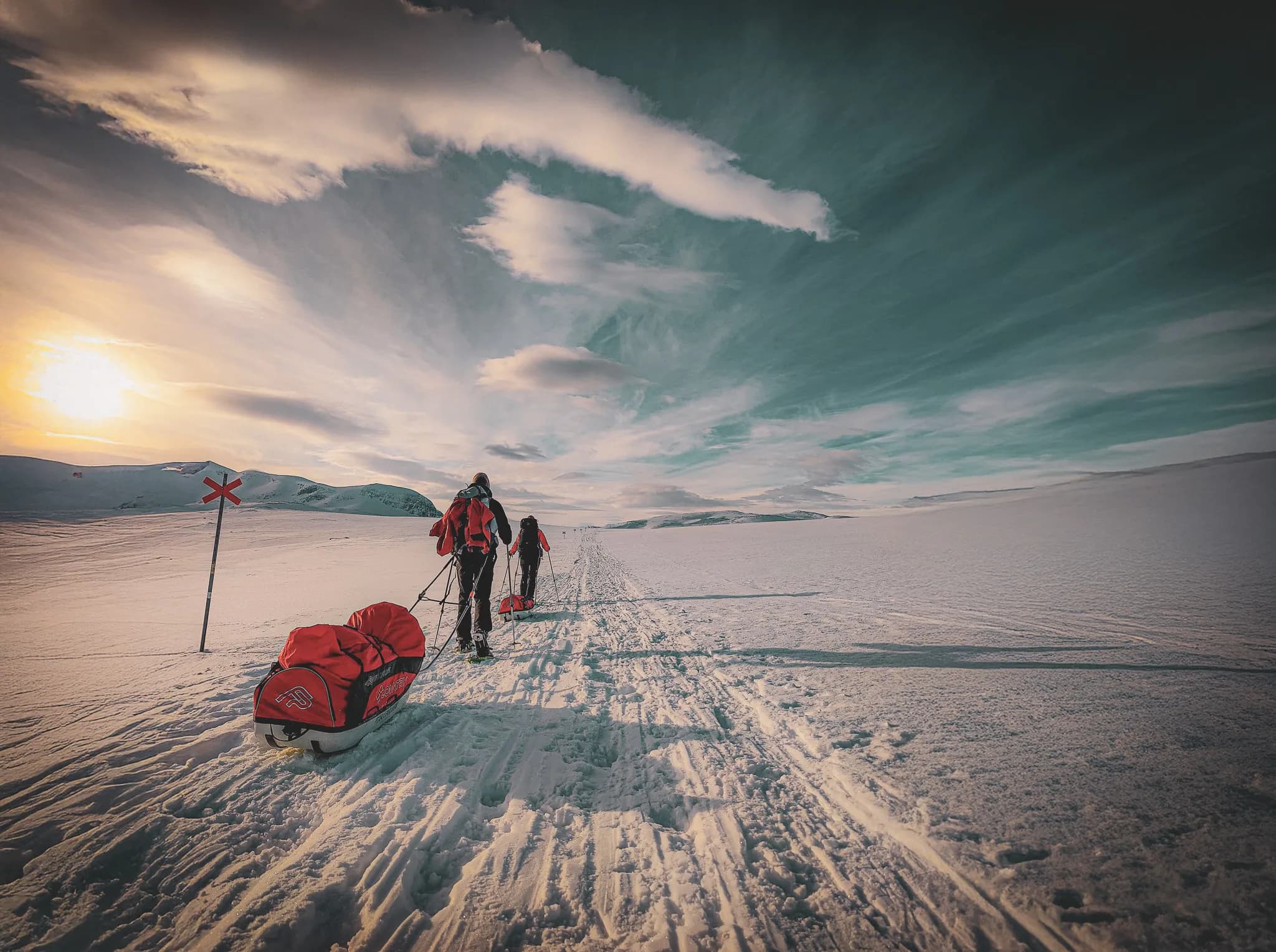 A snow-covered landscape in Swedish Lapland, with hikers pulling red pulkas along a well-marked Kungsleden. The sun is low on the horizon, casting long shadows and creating a warm atmosphere despite the Arctic cold.