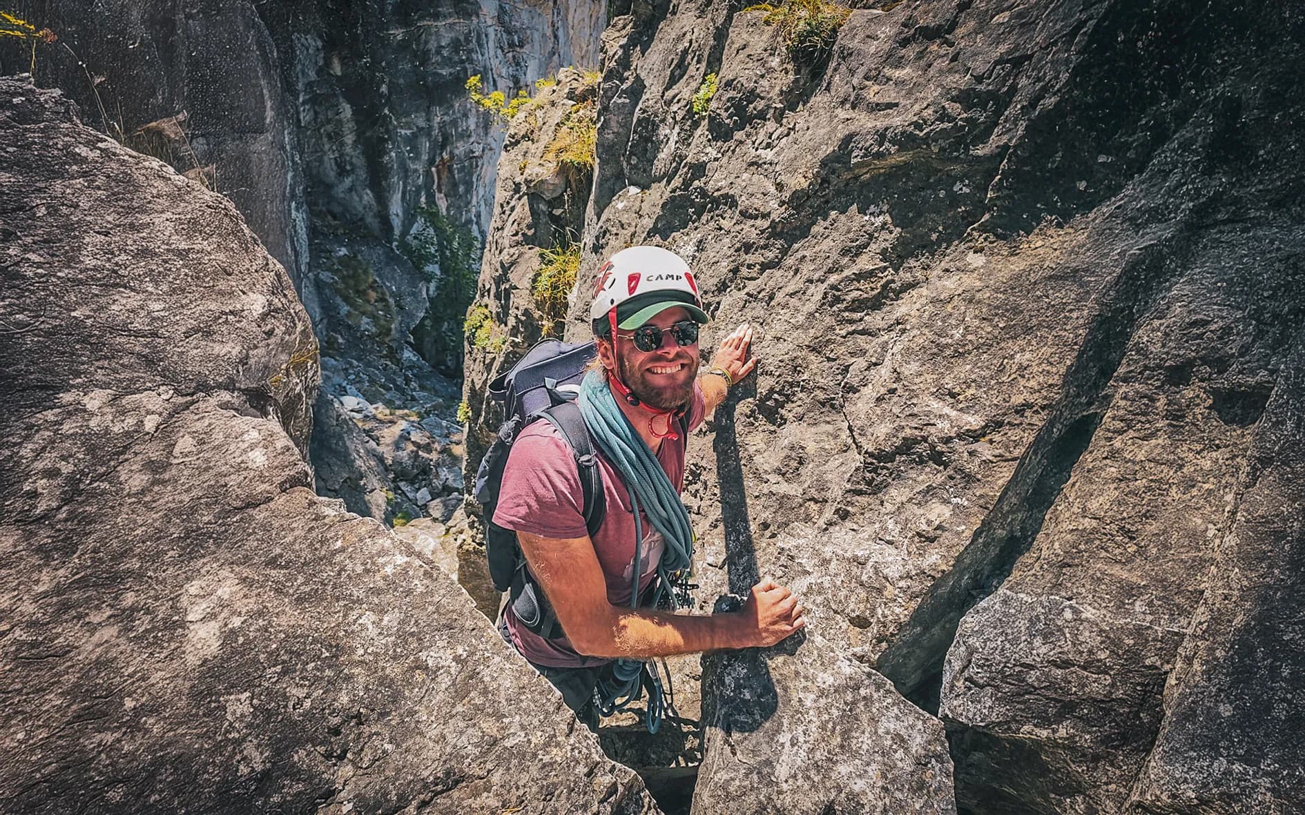 An adventurer climbs a cliff of large grey rocks, surrounded by vertical walls. He is wearing a safety helmet, sunglasses and a T-shirt, with climbing ropes attached.
