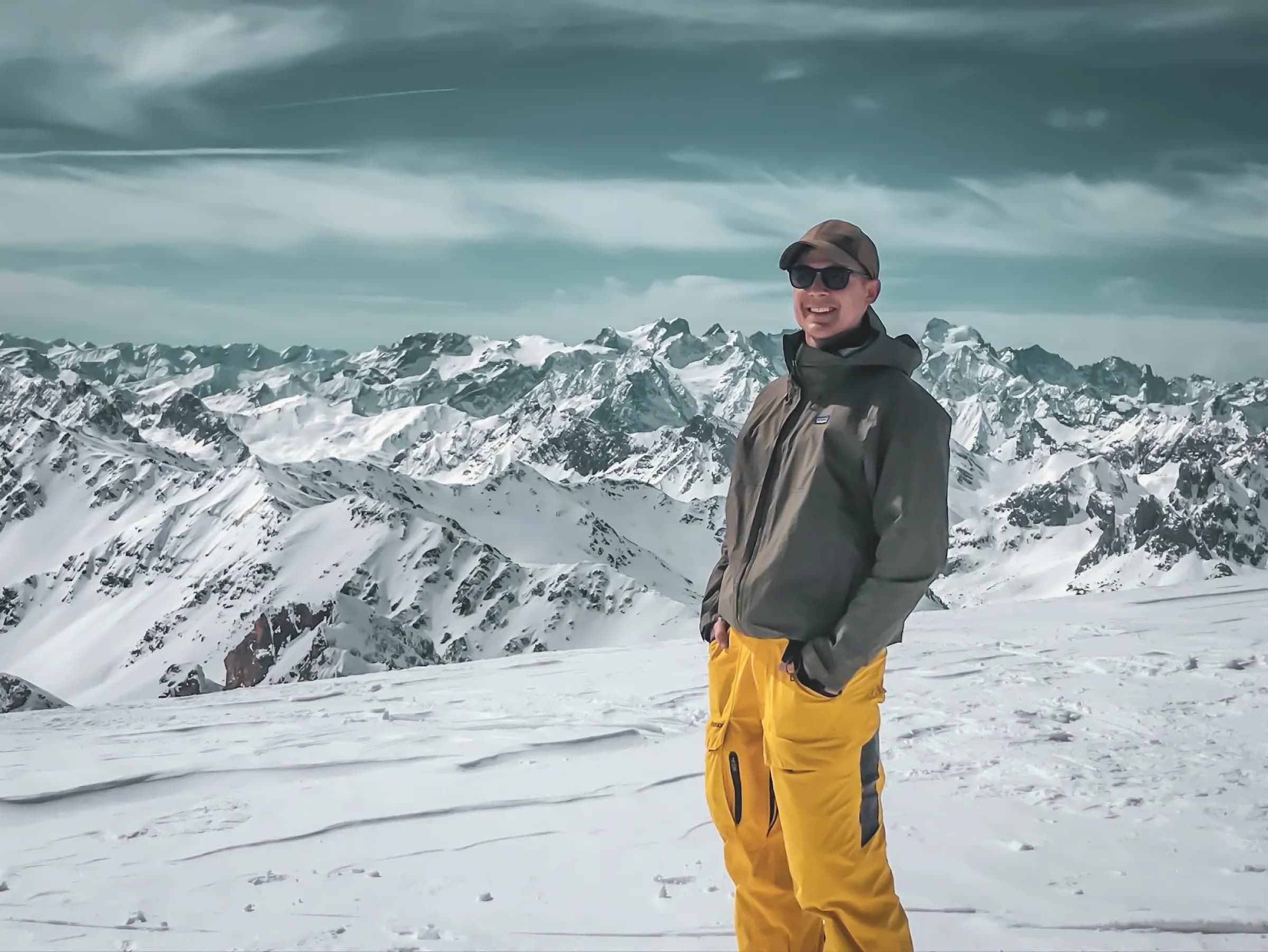 Un homme souriant se tient debout sur un terrain enneigé, entouré de majestueuses montagnes recouvertes de neige. Il porte un manteau gris foncé et un pantalon jaune vif, ainsi qu'une casquette et des