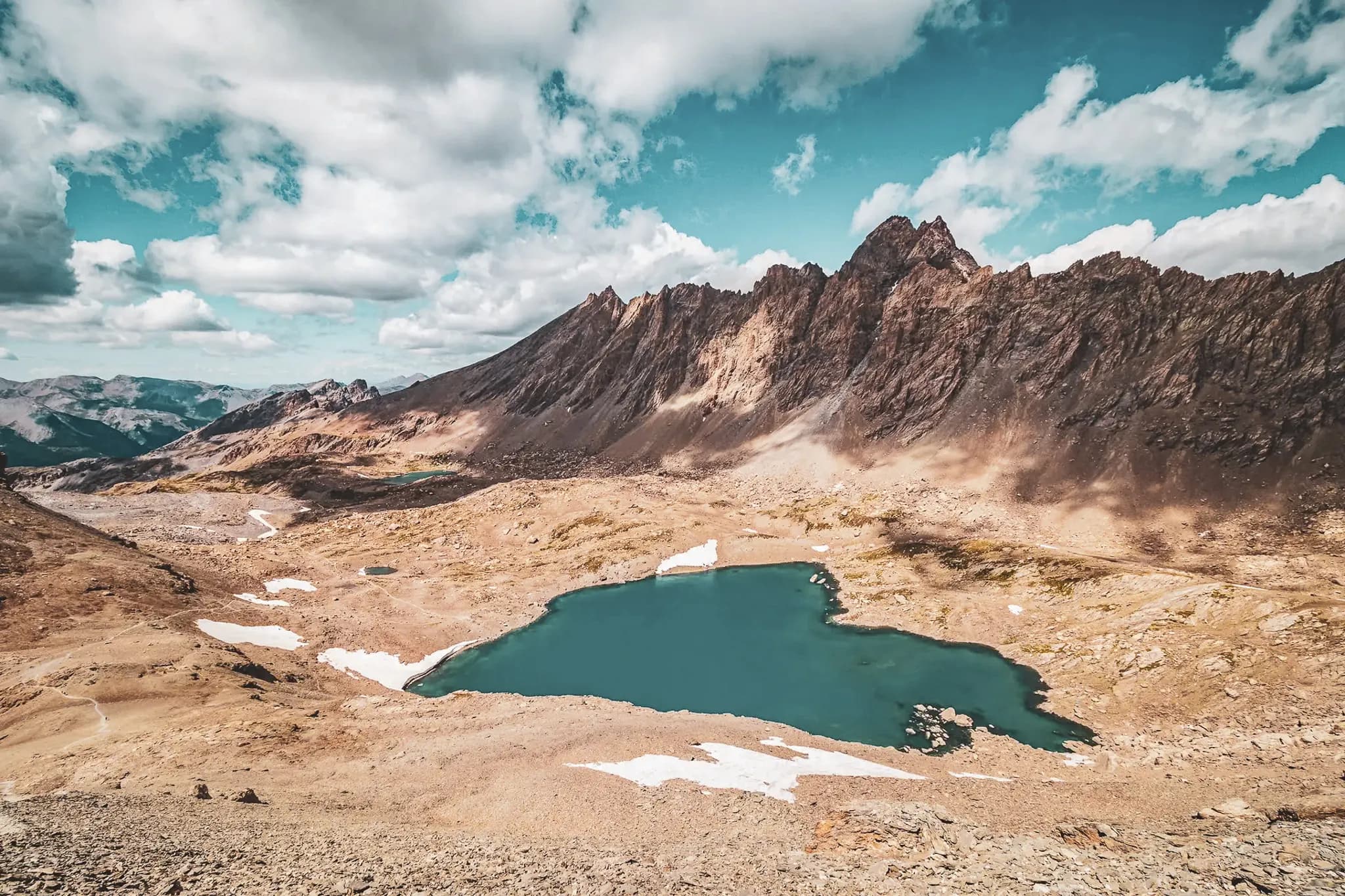A spectacular mountain landscape with rocky peaks in the background, rising up under a blue sky dotted with white clouds. In the foreground, a deep-blue lake, surrounded by arid, rocky terrain.