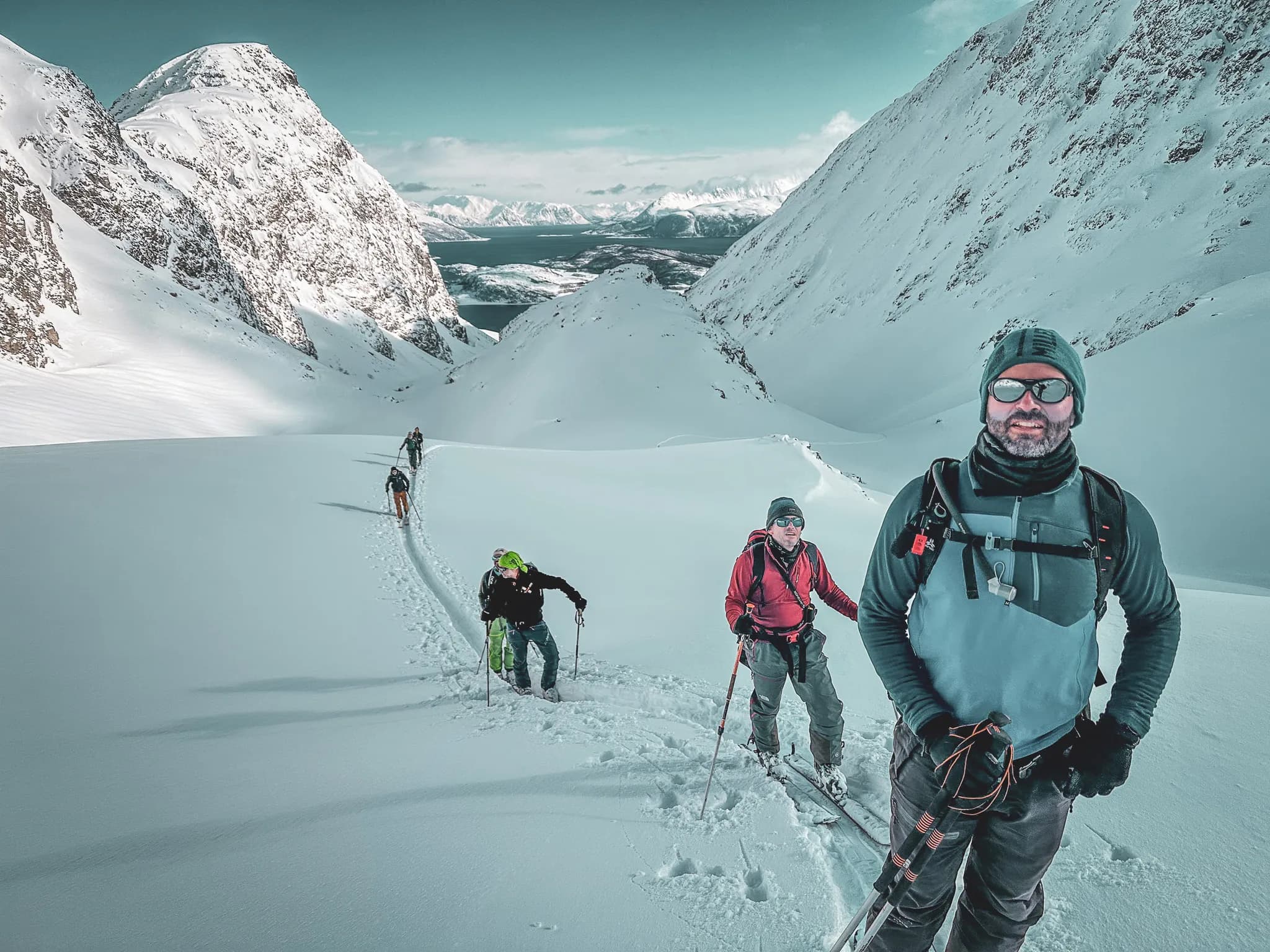 Un groupe de quatre alpinistes progresse sur un vaste terrain neigeux, entouré de montagnes majestueuses et enneigées. L'un d'eux, situé au premier plan, porte des lunettes de soleil, un bonnet et une veste