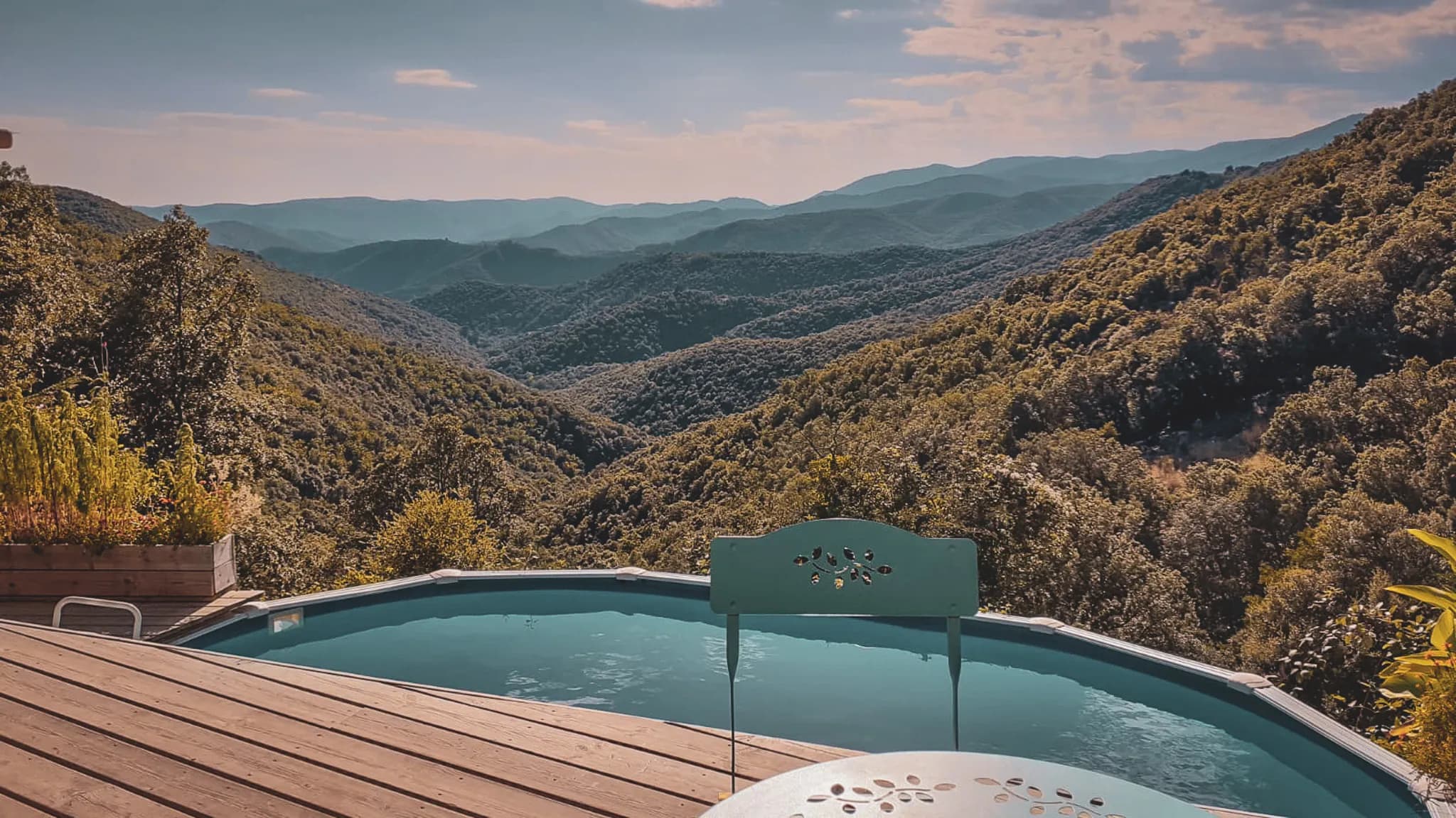 A panoramic view of green hills, with a swimming pool in the foreground. The hexagonal-shaped pool reflects the sky and the surrounding mountains. Next to it, a round table and a chair with a
