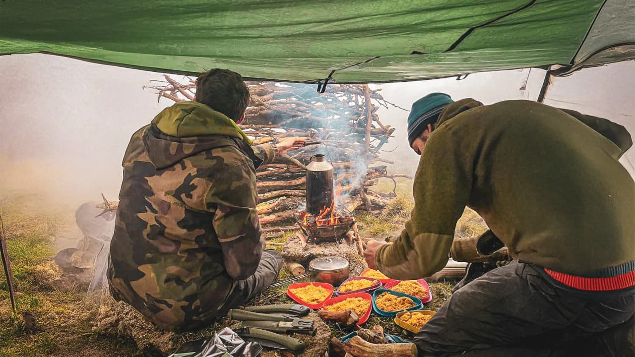 Two adventurers warm themselves by the campfire in a green tent, a colourful meal spread out on the ground.