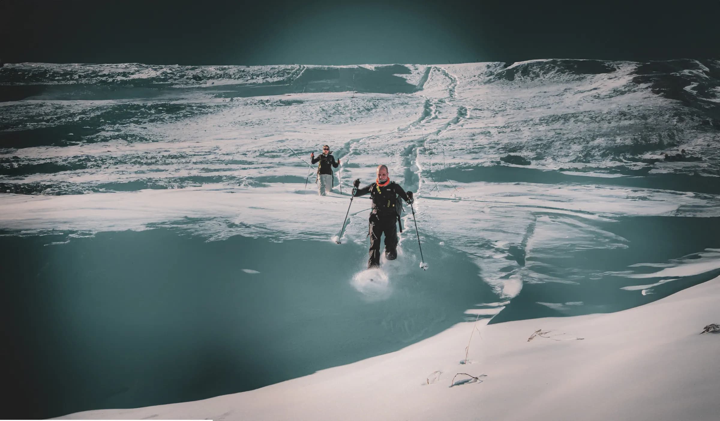 A snowy landscape with two skiers crossing an expanse of snow. One of the skiers, in the foreground, is walking with poles, leaving tracks in the untouched snow. In the background, the other skier