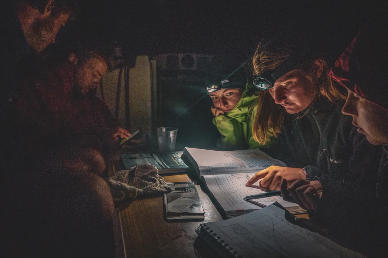 Een groep jongeren op een boot, gefascineerd door zeekaarten, in gedempt licht.